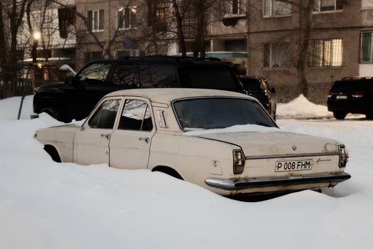 An old car partially buried in snow on a city street during winter.