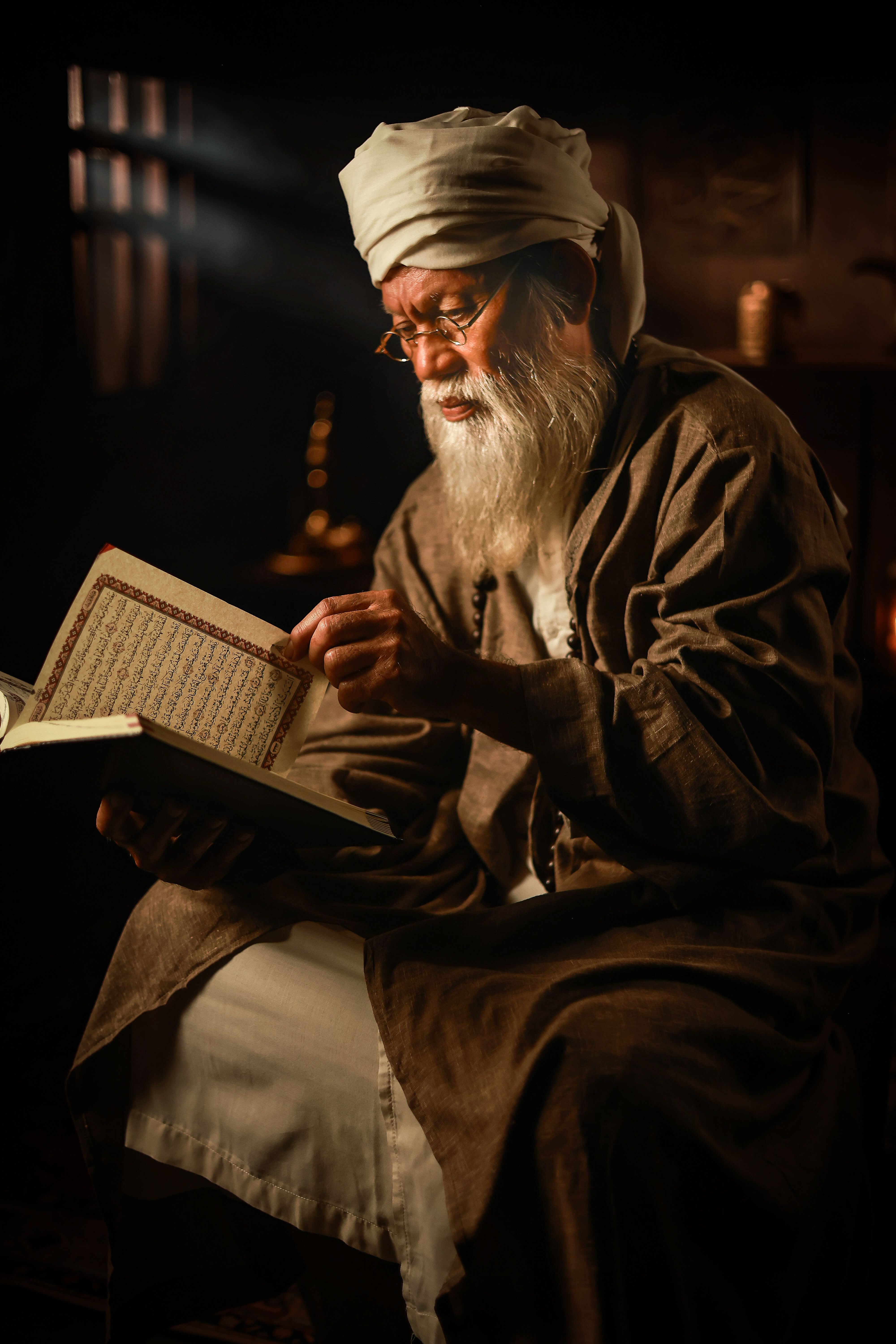 An elderly man wearing traditional attire reads an ancient book under soft, focused lighting in a dimly lit room.