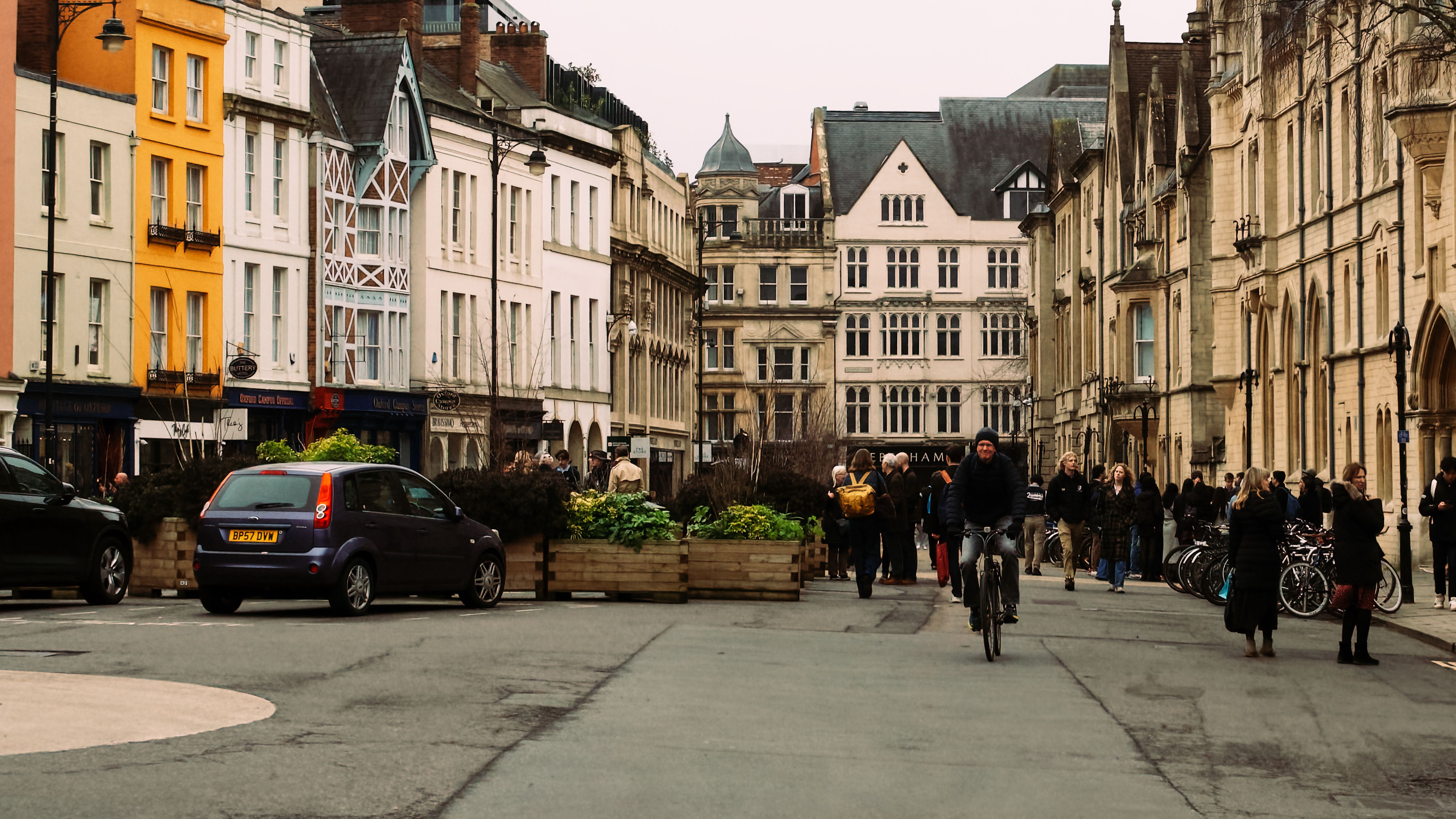 A lively day on an Oxford street with people and a cyclist amidst historic architecture.