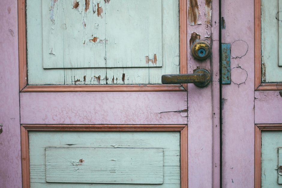 Close-up image of a vintage wooden door with peeling pastel paint and a rusted handle.