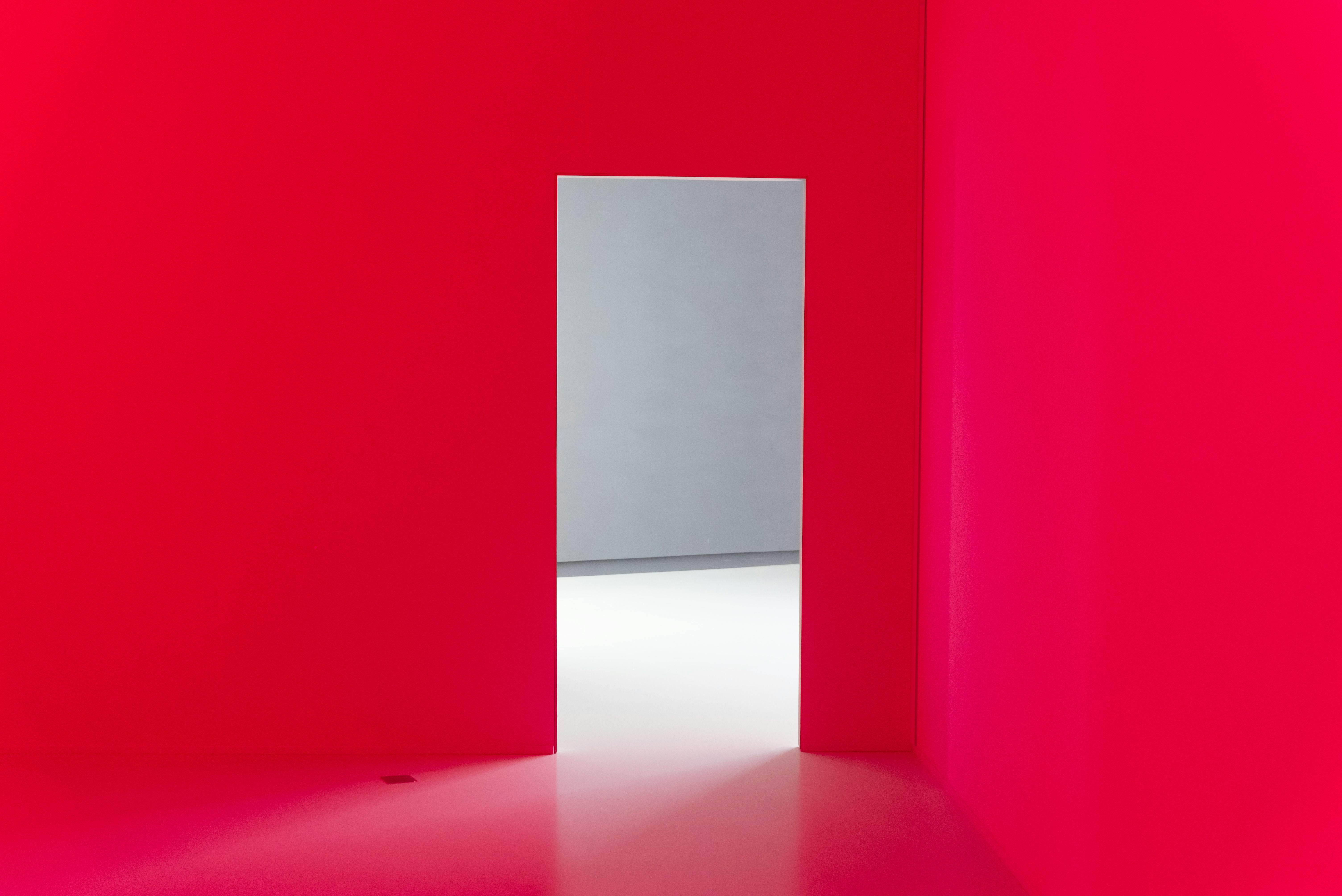 Free Abstract interior with bright red walls and an open doorway leading to a minimalist white room. Stock Photo