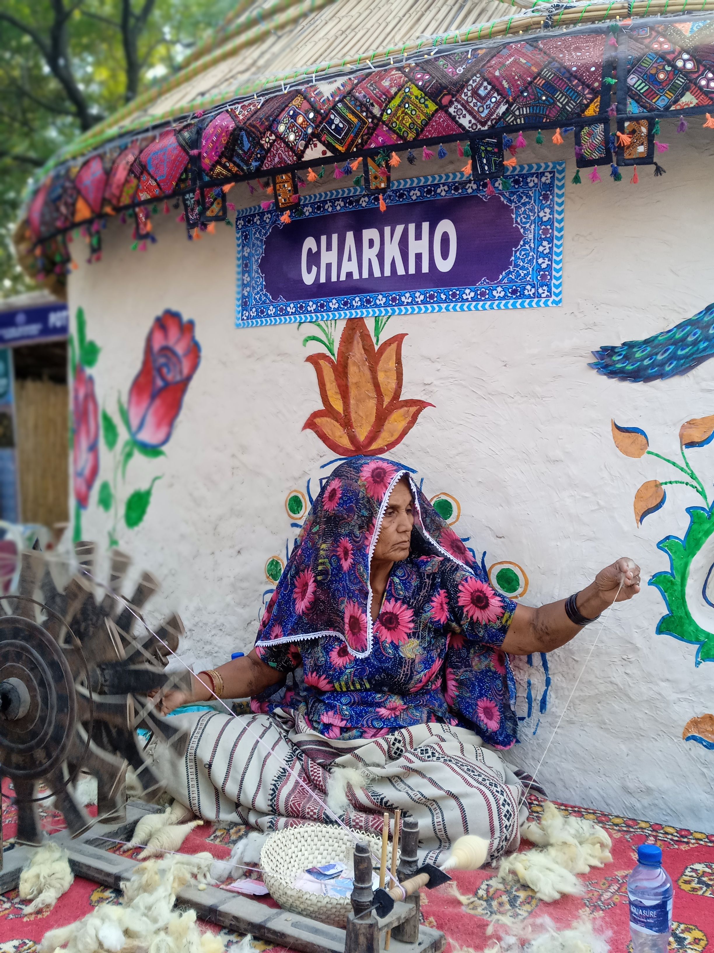 A South Asian woman spins yarn using a traditional charkho in a vibrant, artistic setting.