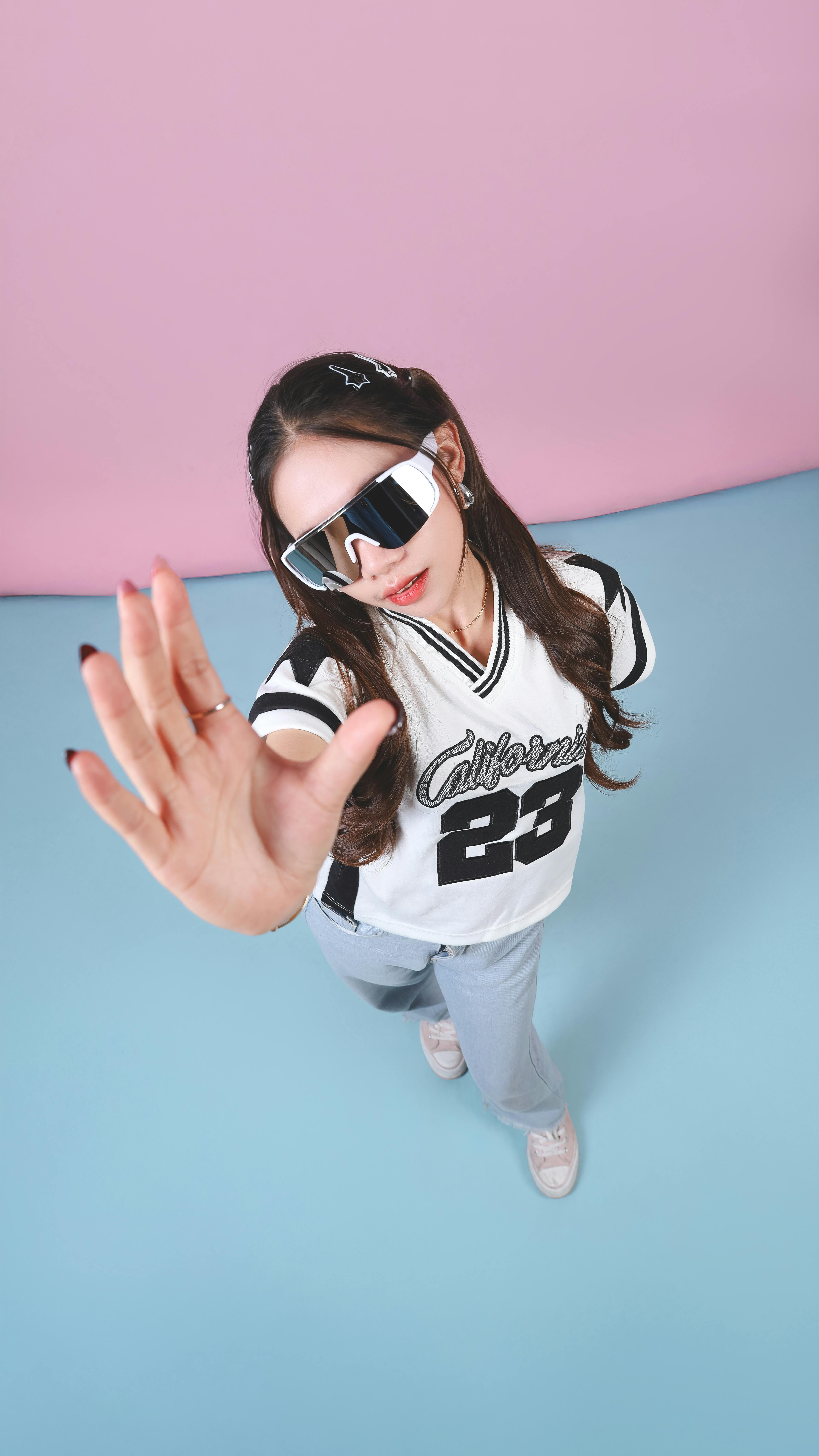 無料 パステル調の背景にスポーツジャージとサングラスを着けてスタイリッシュにポーズをとる若い女性。 写真素材