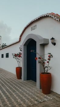 A quaint white house with a blue door and red tiled roof, featuring potted flowering plants and lanterns.