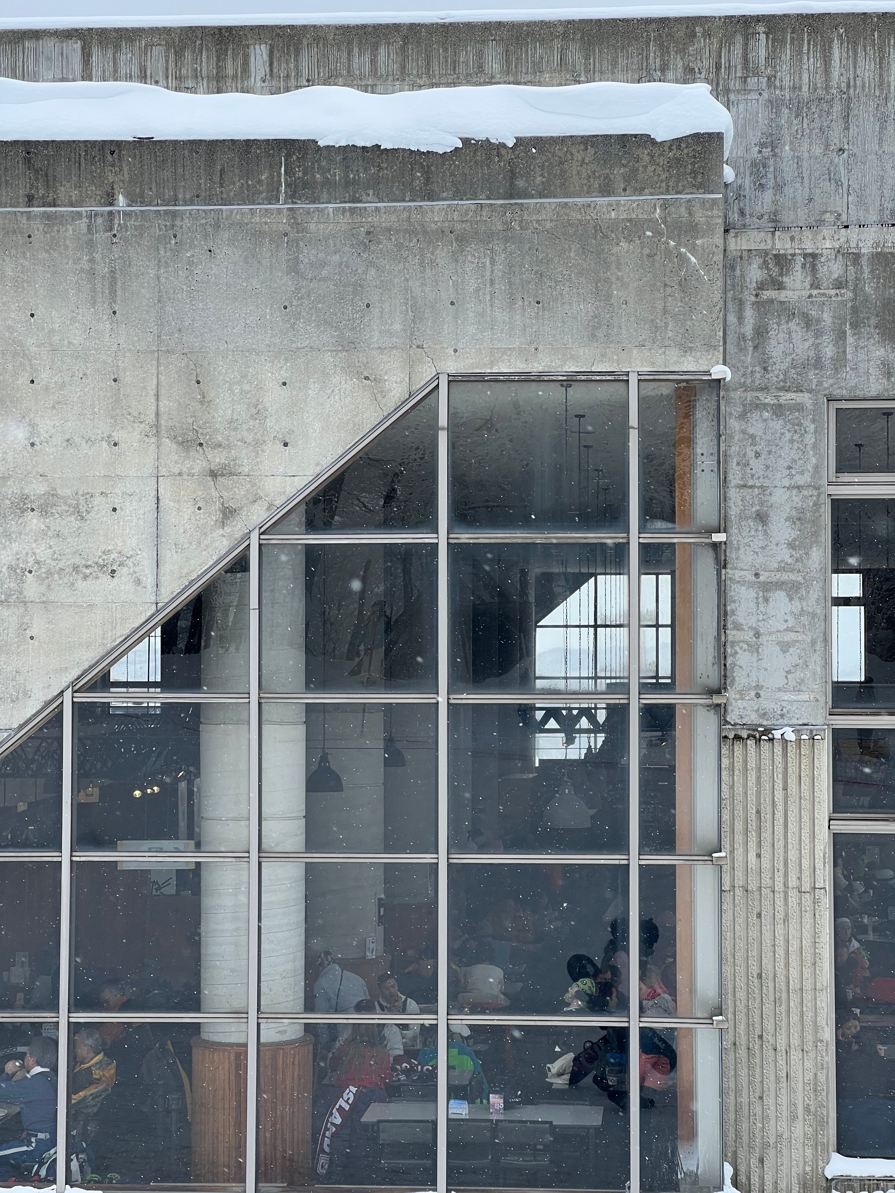Skiers at tables behind a tall window in a concrete building, with snow on the roof and snow falling