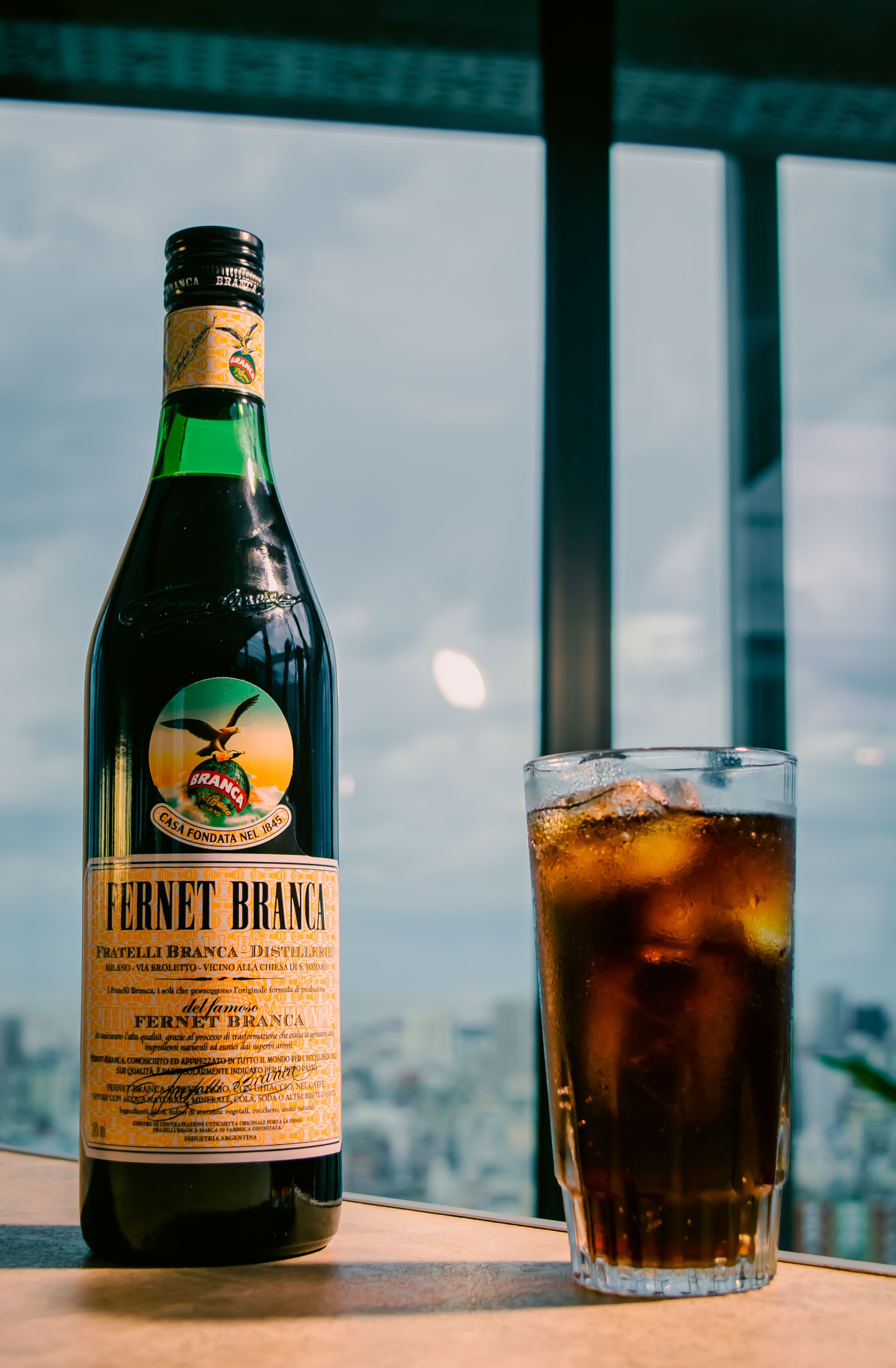 Fernet Branca bottle and glass with drink placed on a table against a cityscape backdrop.