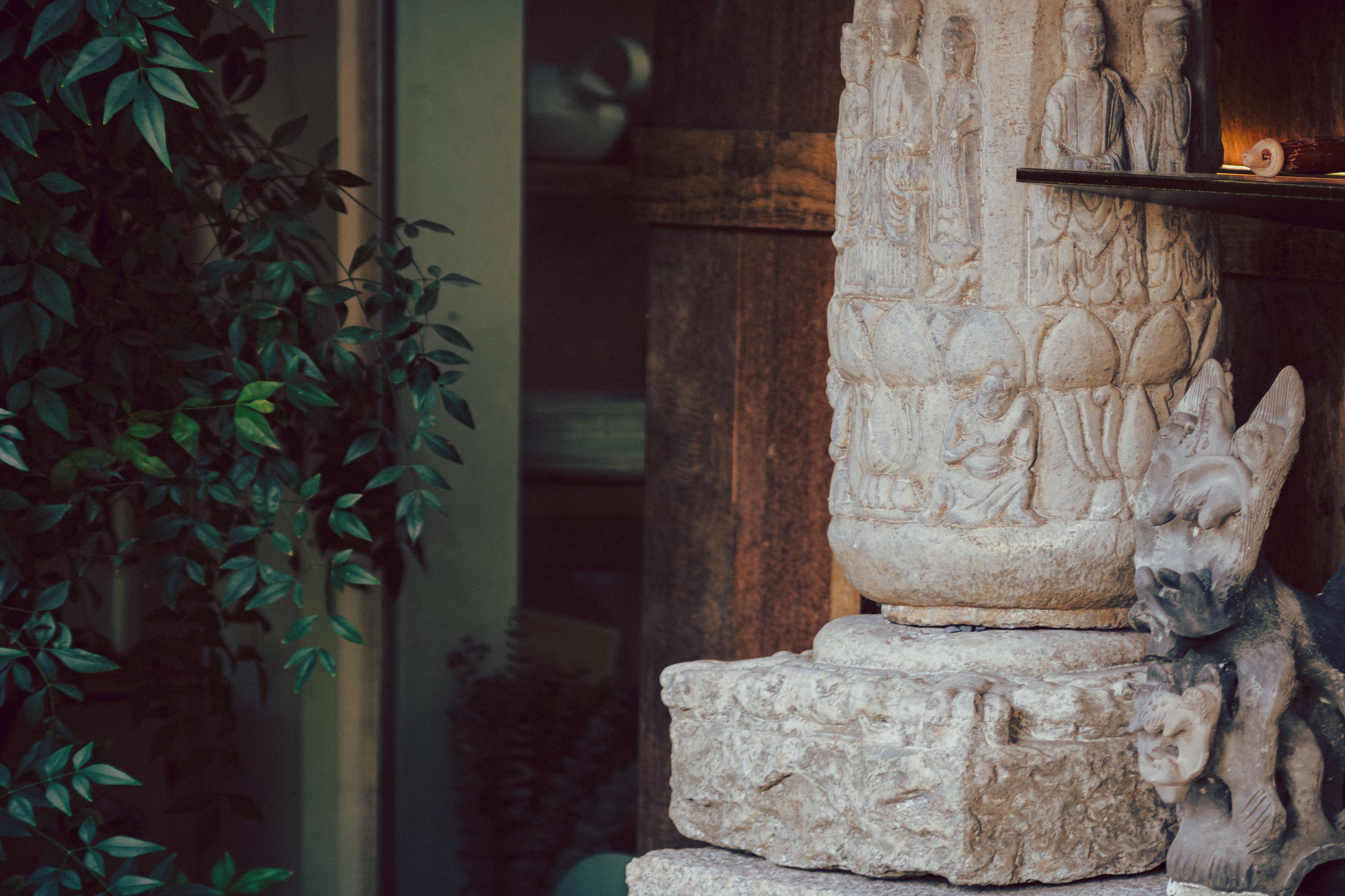 Intricate stone sculpture with carved figures surrounded by lush greenery indoors.