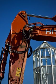 Close-up of an excavator arm with a glass structure in the background and clear blue sky.