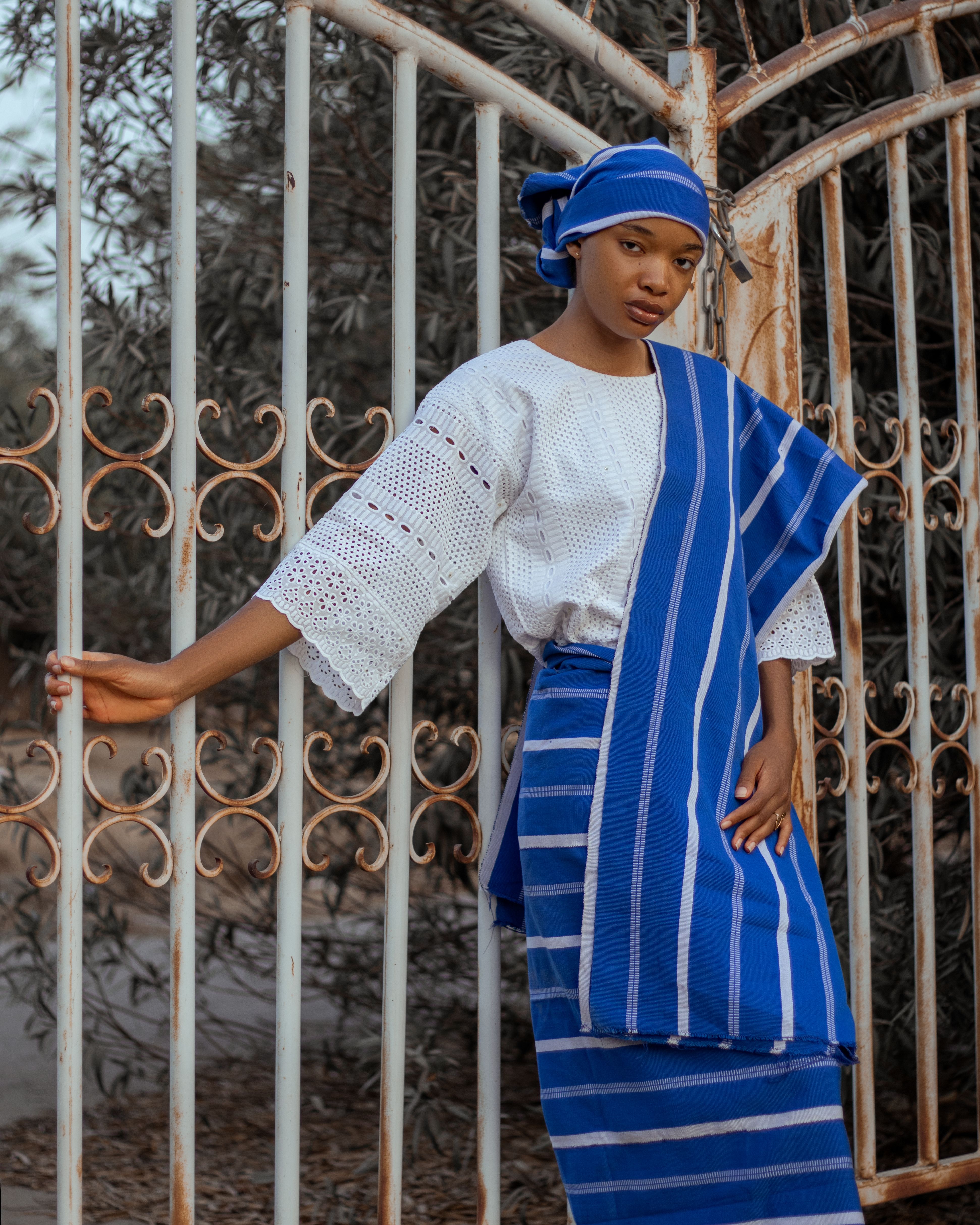 A young woman in traditional Nigerian clothing posing outdoors by an iron gate.
