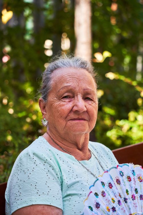 Free Elderly woman sitting on a bench with a decorative fan in a sunny, green park. Stock Photo