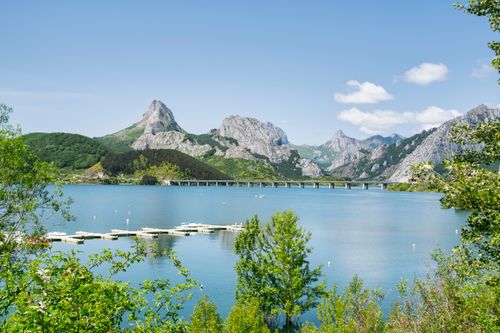 Free Beautiful view of a mountain lake with a bridge, surrounded by lush greenery in summer, under a bright blue sky. Stock Photo