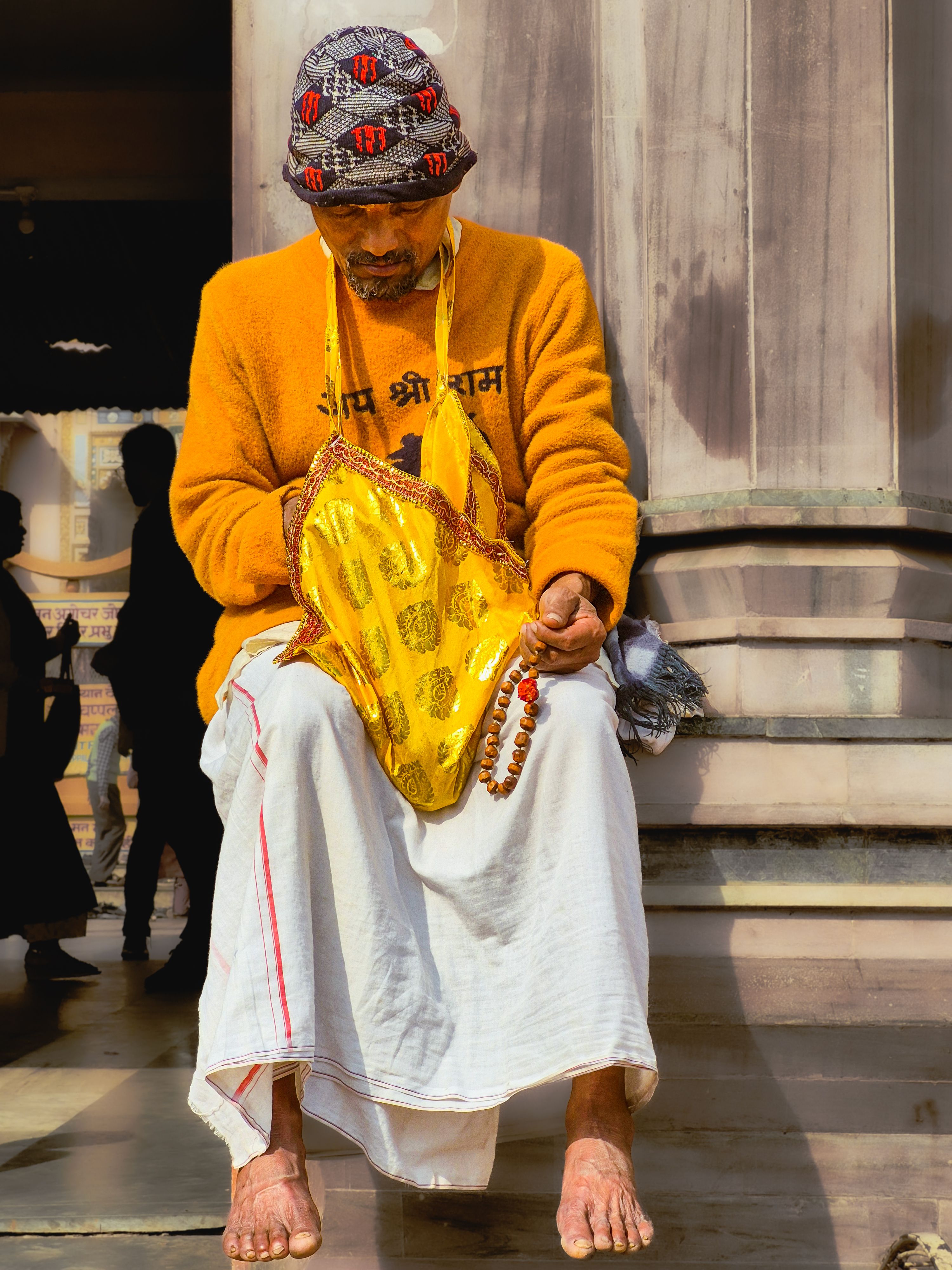 An elderly man in traditional attire prays with beads outside a temple.