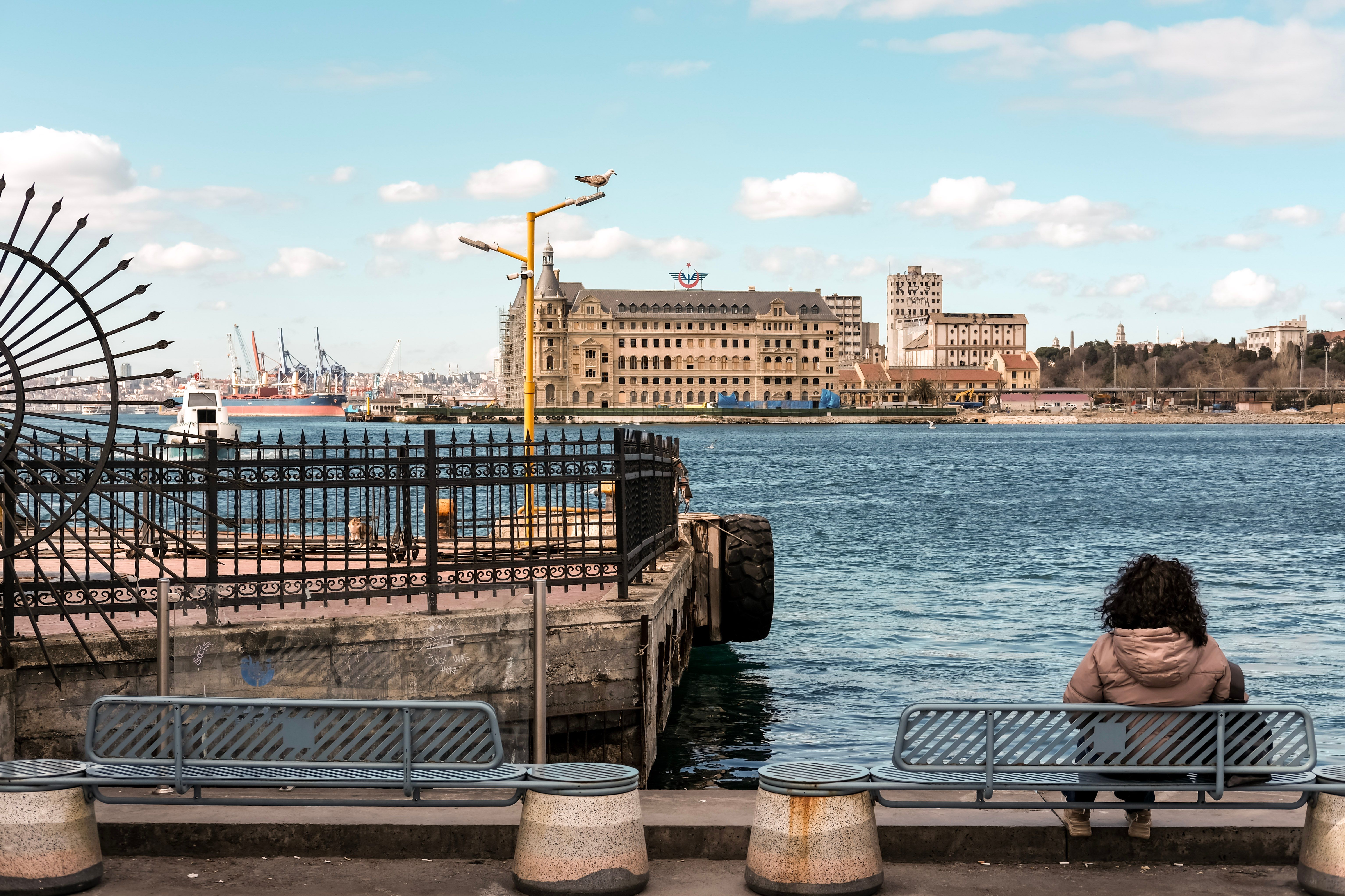 Woman sitting by Istanbul waterfront with Haydarpaşa Train Station in background, offering a serene urban landscape.