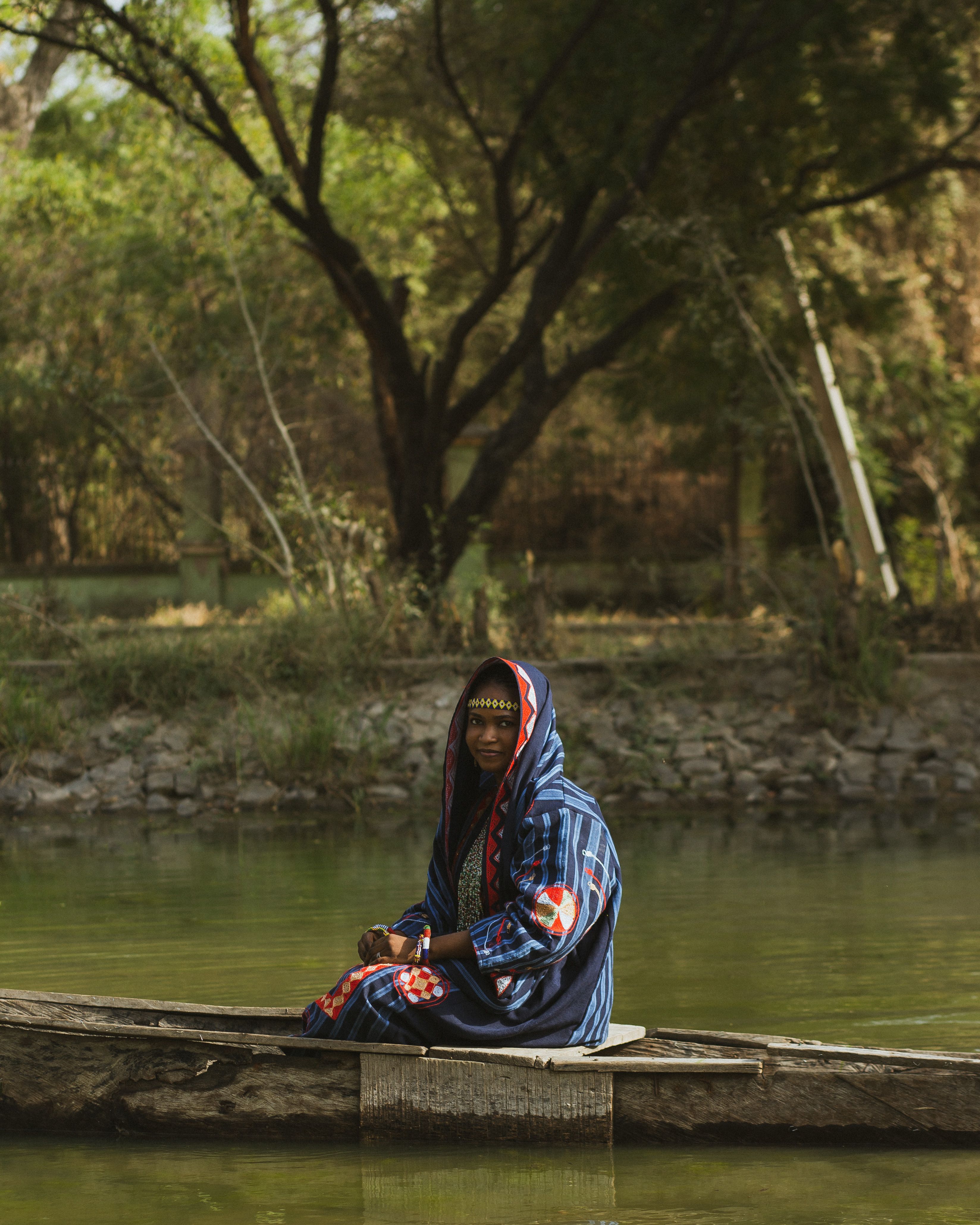 A Nigerian woman wearing traditional clothing sits on a canoe in a serene outdoor setting.