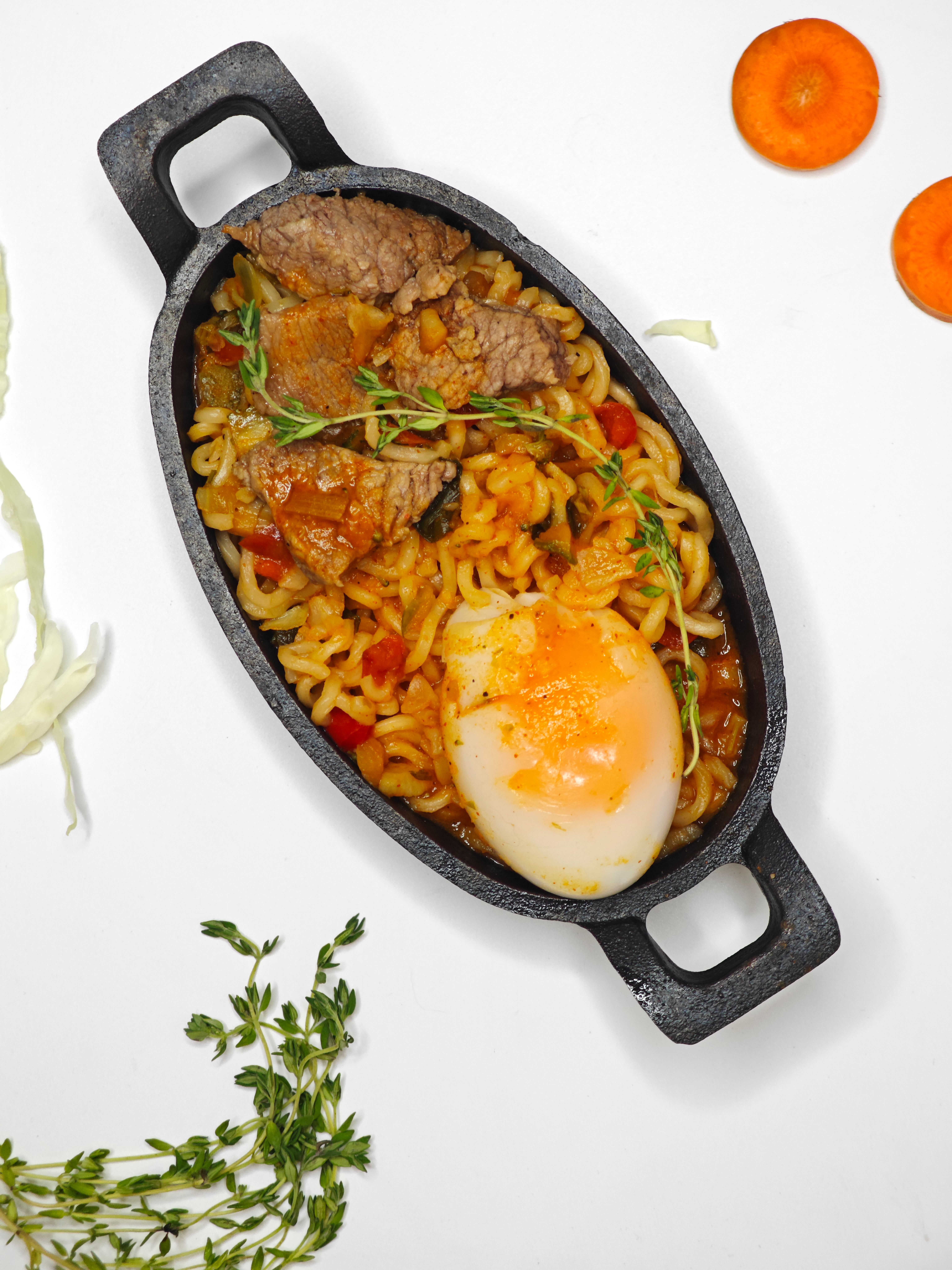 Appetizing ramen with beef and a soft-boiled egg in a black skillet, garnished with herbs.
