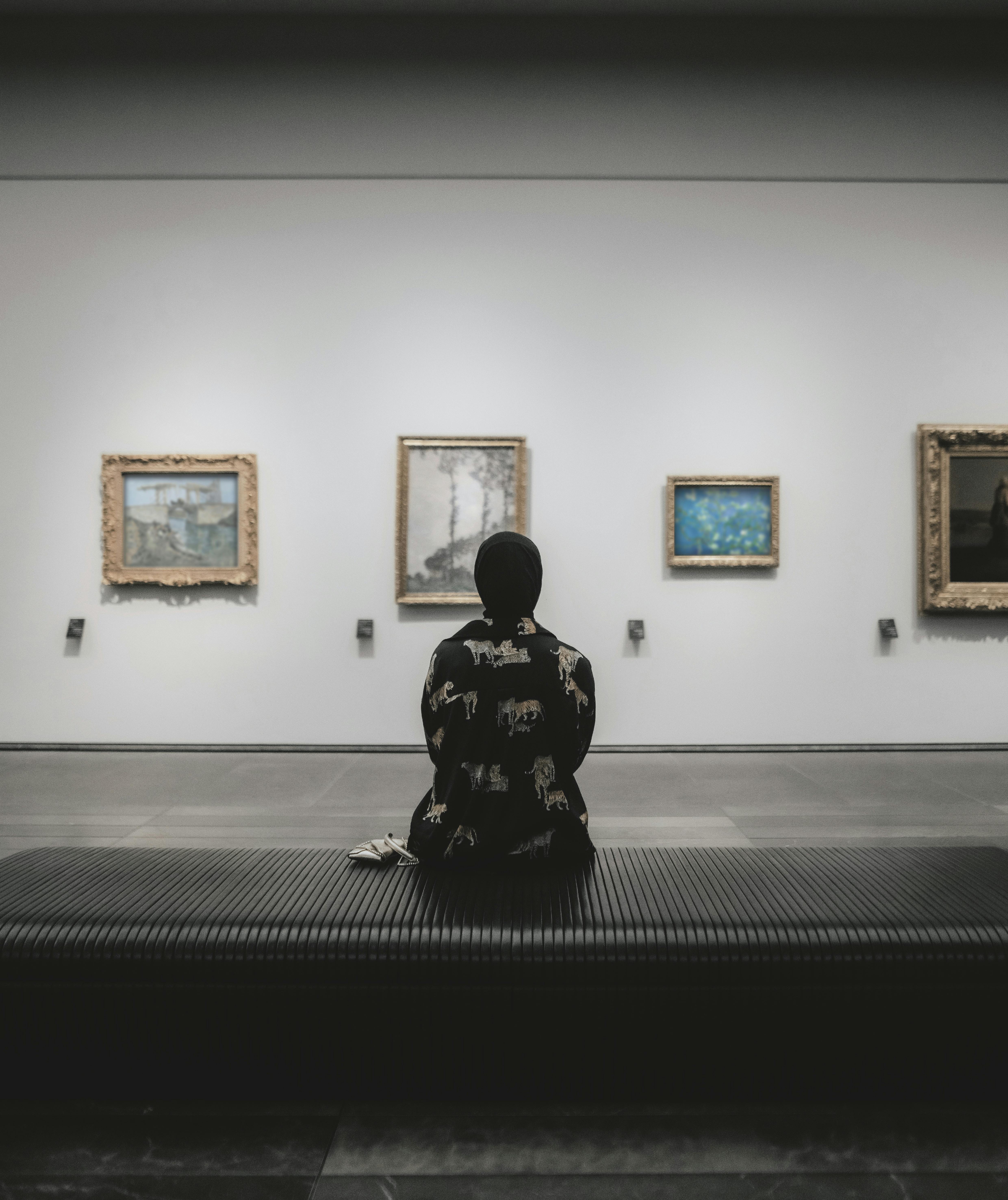 Gratuit Une personne assise sur un banc dans une galerie d'art, contemplant des tableaux. Photos
