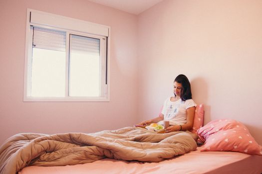 A woman relaxes with breakfast in bed in a cozy pink room, enjoying a peaceful morning.