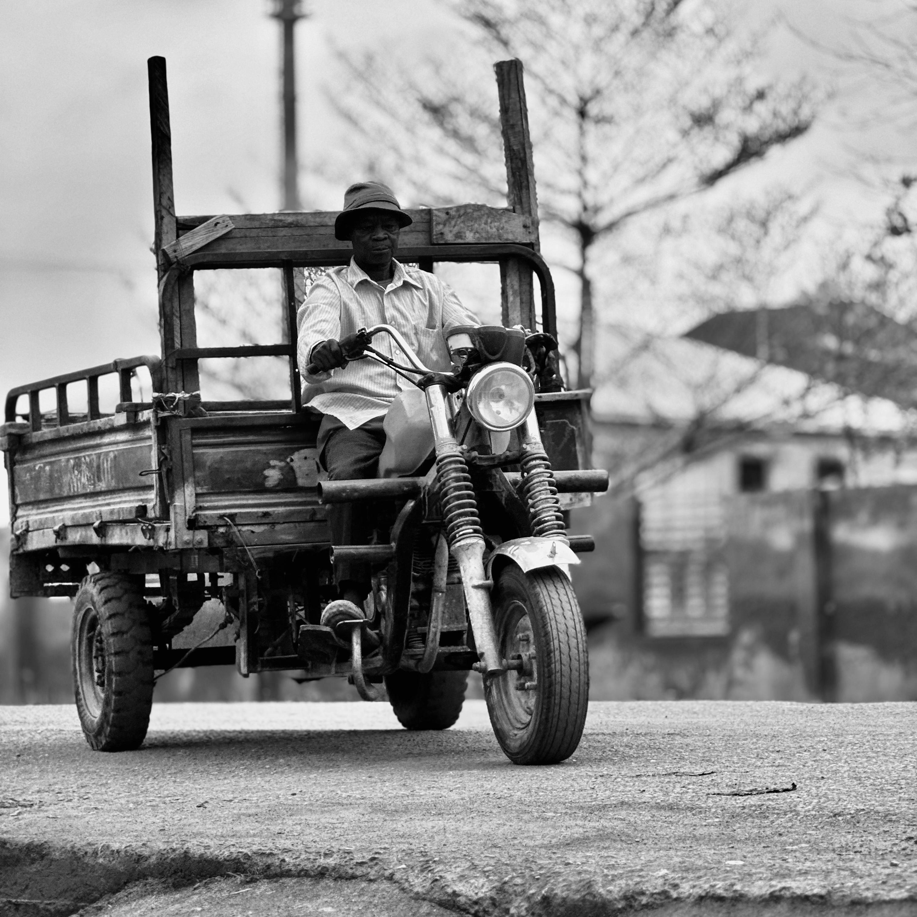Black-and-white photo of a man riding a tricycle motorbike on a street in Accra, Ghana.
