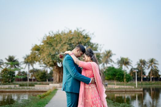 A couple in traditional attire embraces lovingly beside a tranquil pond with palm trees.