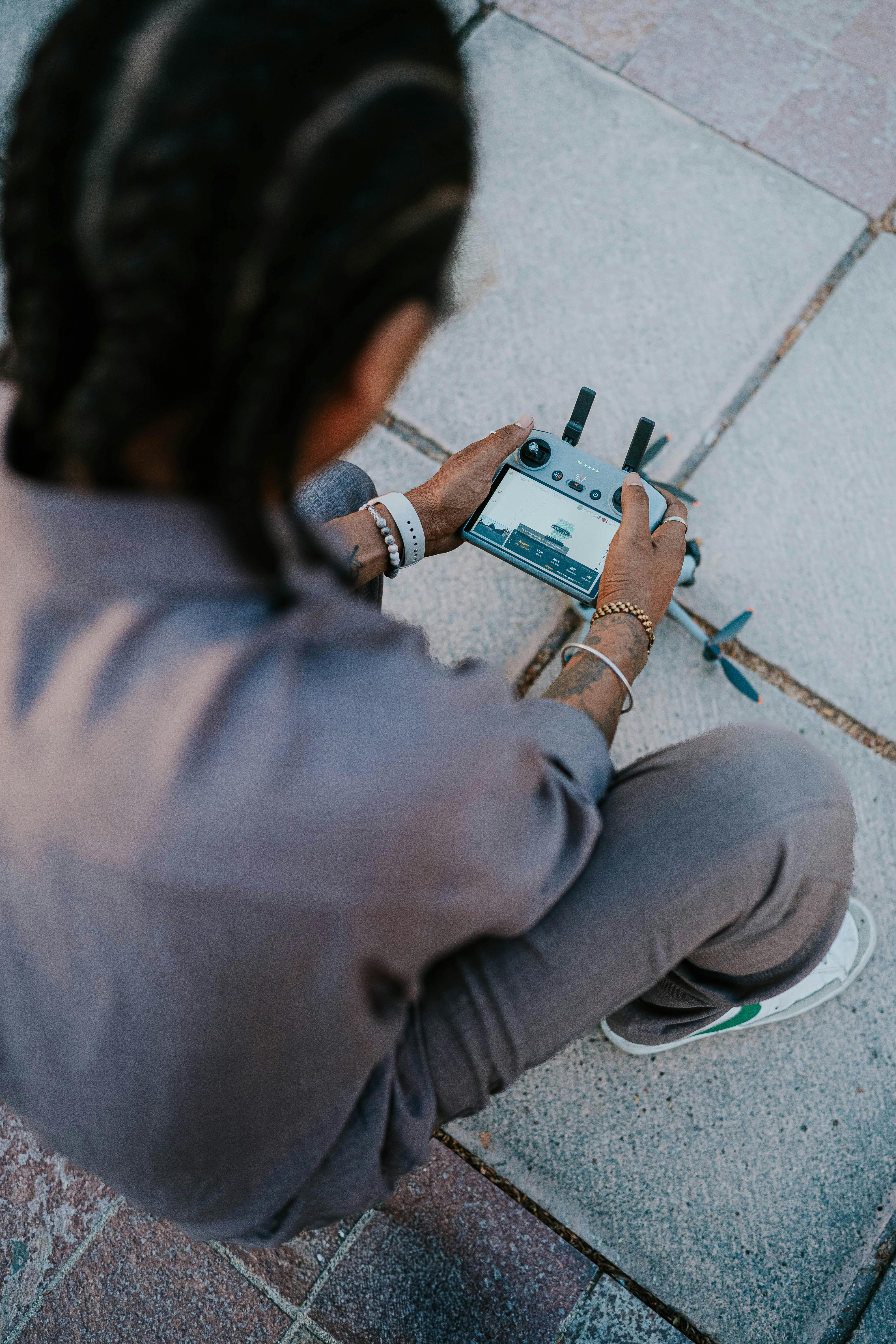 Free A young individual controlling a drone with a remote, seated on paved ground outdoors. Stock Photo