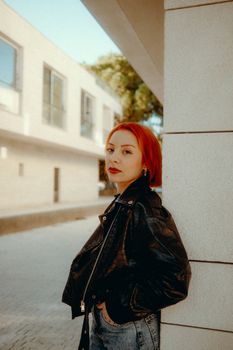 Confident woman with red hair poses in a trendy leather jacket against an urban backdrop.