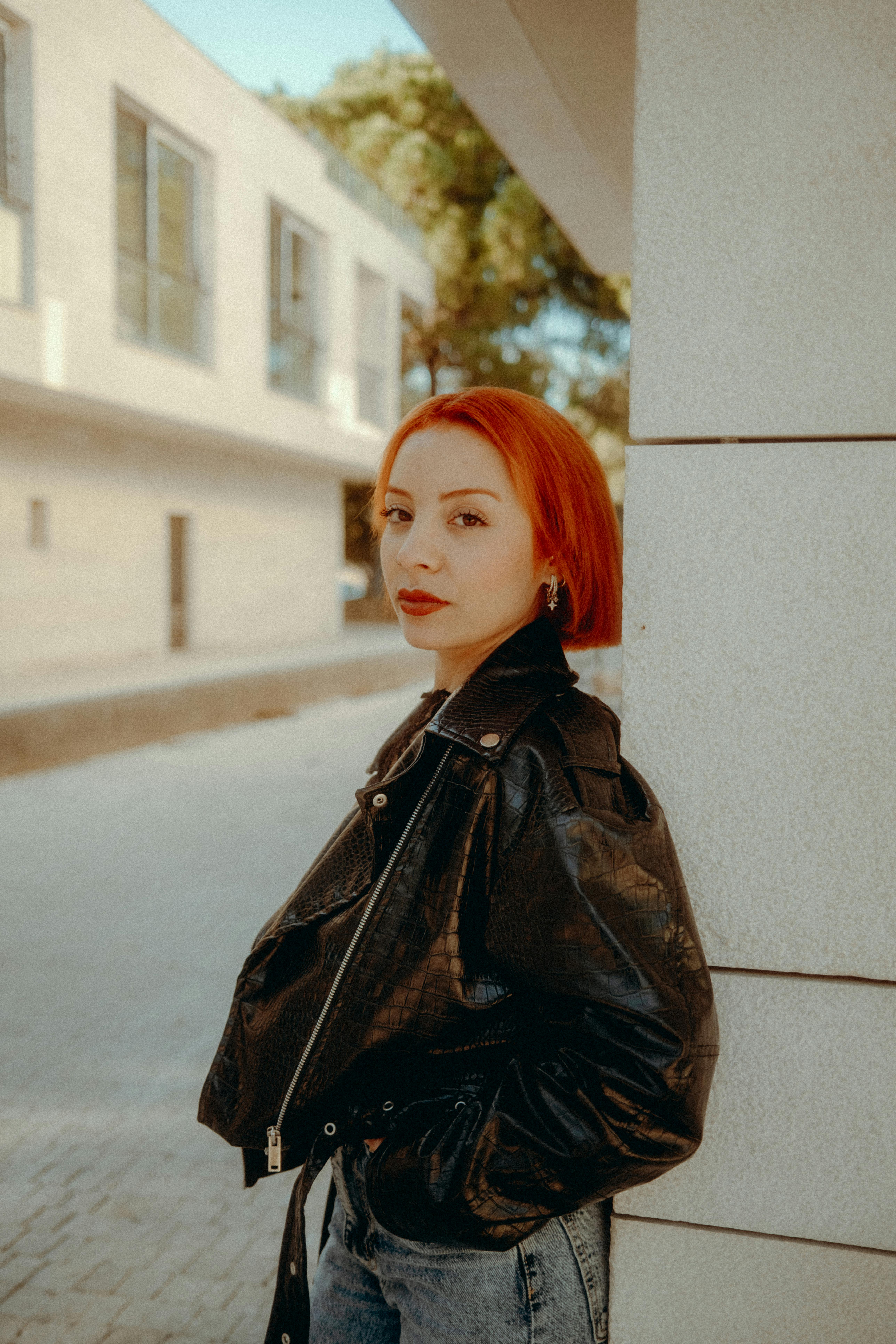 Stylish red-haired woman leans against a wall in an urban setting. Modern and fashionable.