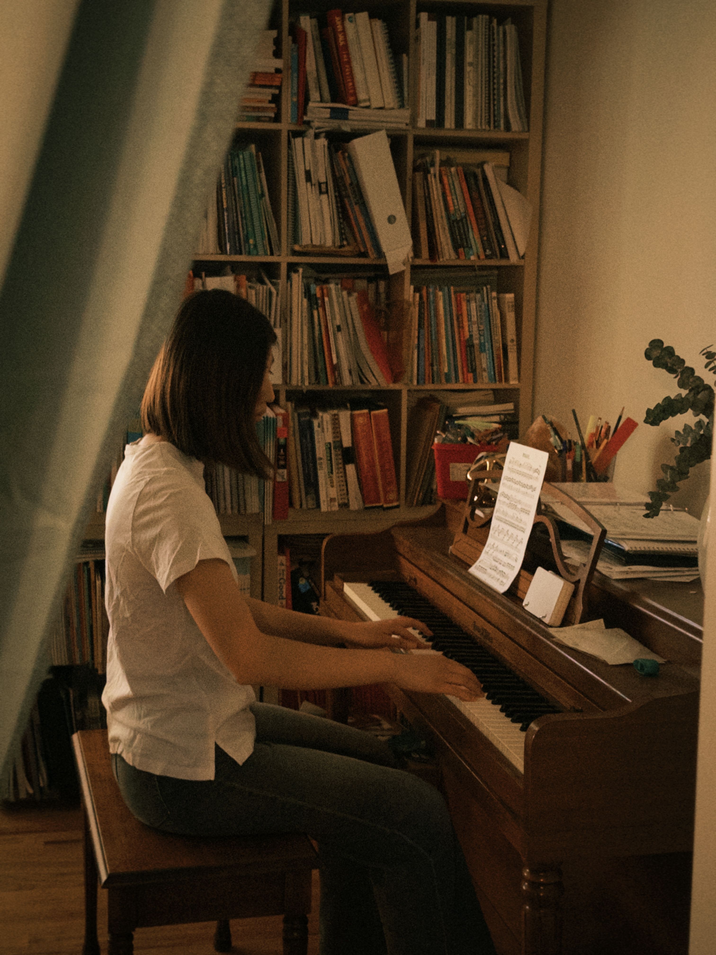 A woman plays the piano in a cozy room, surrounded by a bookshelf filled with books and illuminated by soft lighting.