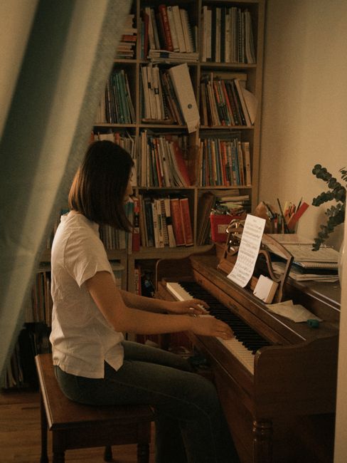 A woman plays the piano in a cozy room, surrounded by a bookshelf filled with books and illuminated by soft lighting.