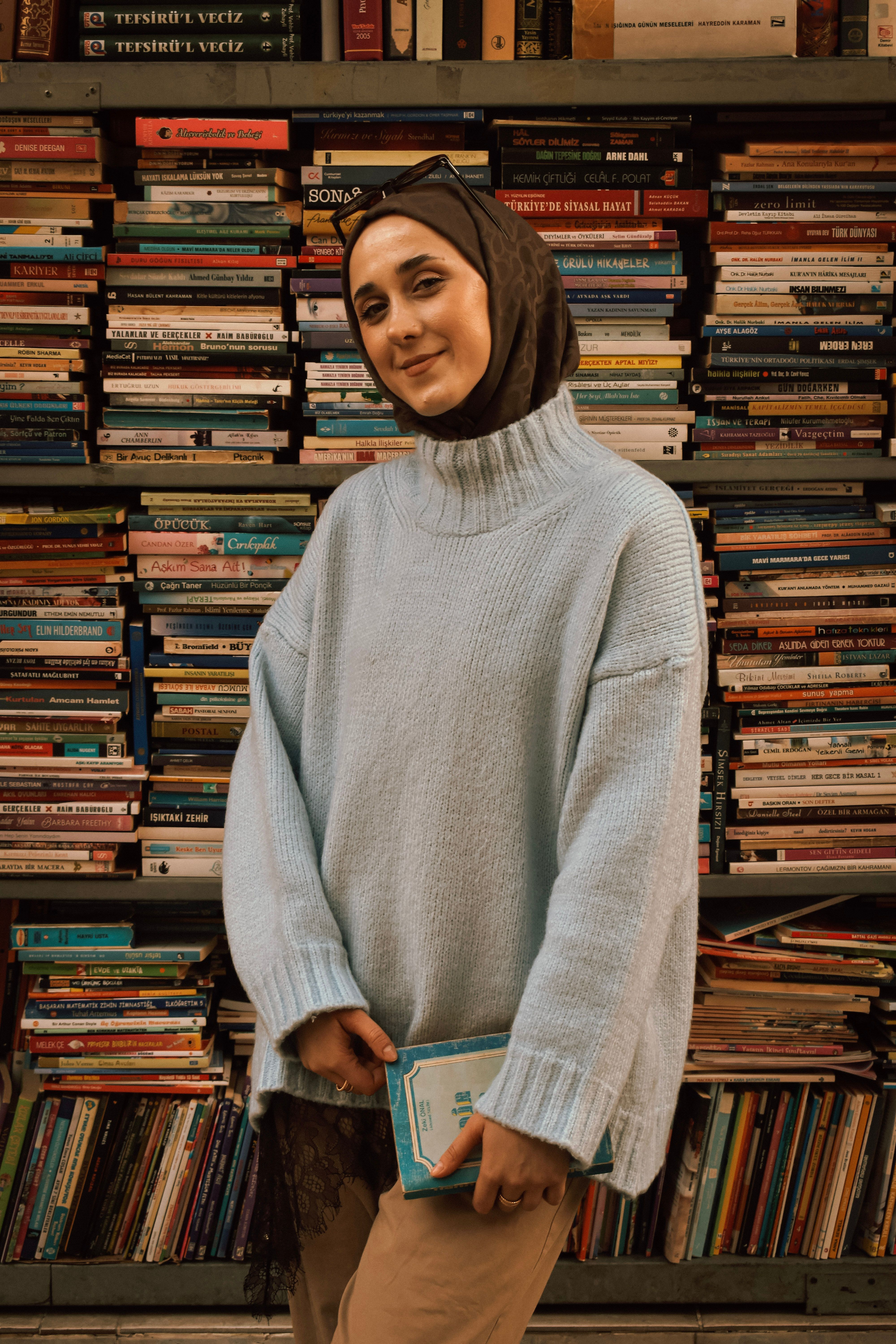A woman in a cozy sweater stands in front of a bookshelf in a Bursa bookstore, Türkiye.