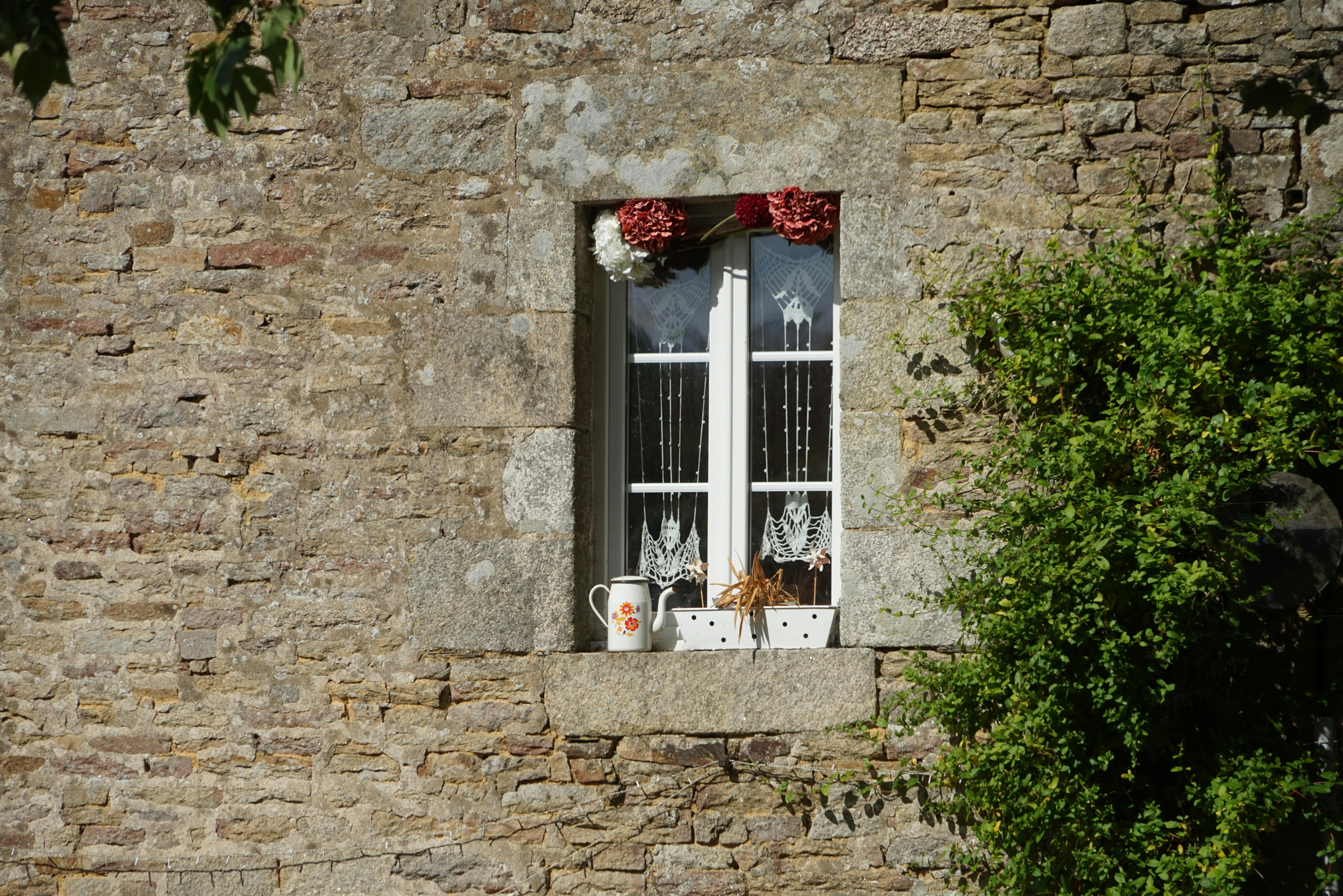 Rustic stone house window adorned with cotton curtains and plants.