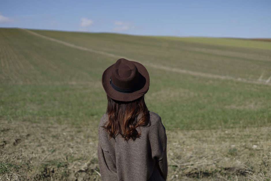 A woman wearing a brown hat looks at an expansive open field under a clear sky.