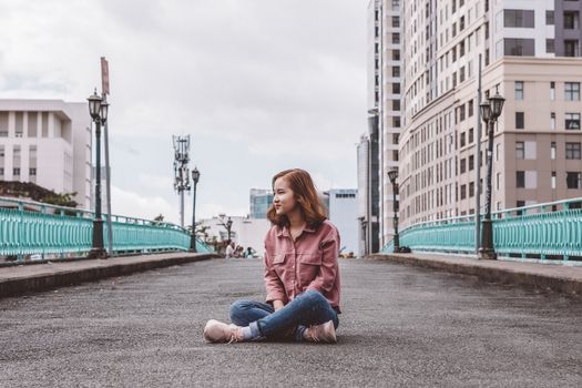 一名年轻女子穿着休闲装，坐在城市的一座桥上，微笑着移开目光