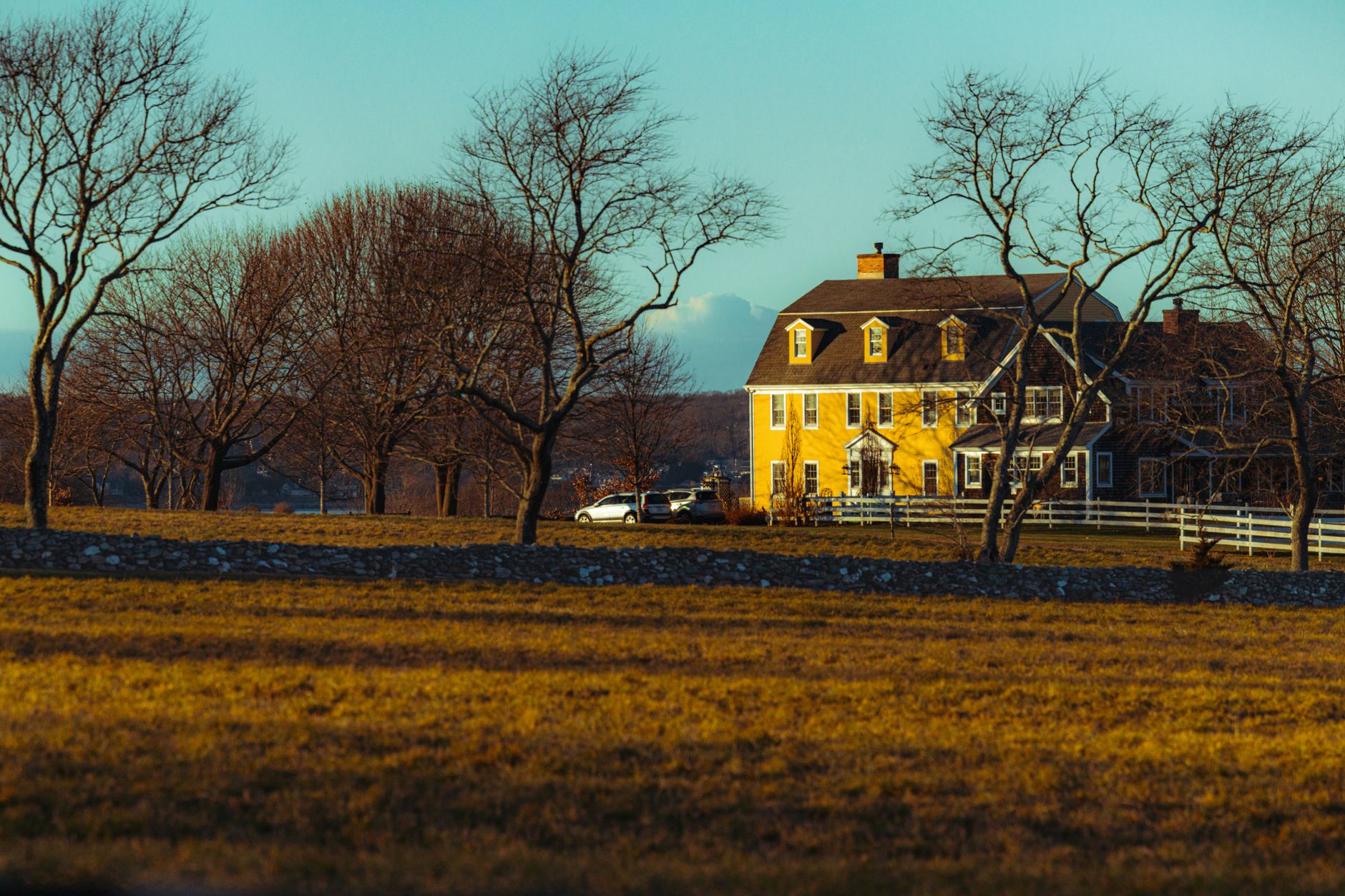 A picturesque yellow country house amidst bare trees during golden hour in rural New England.