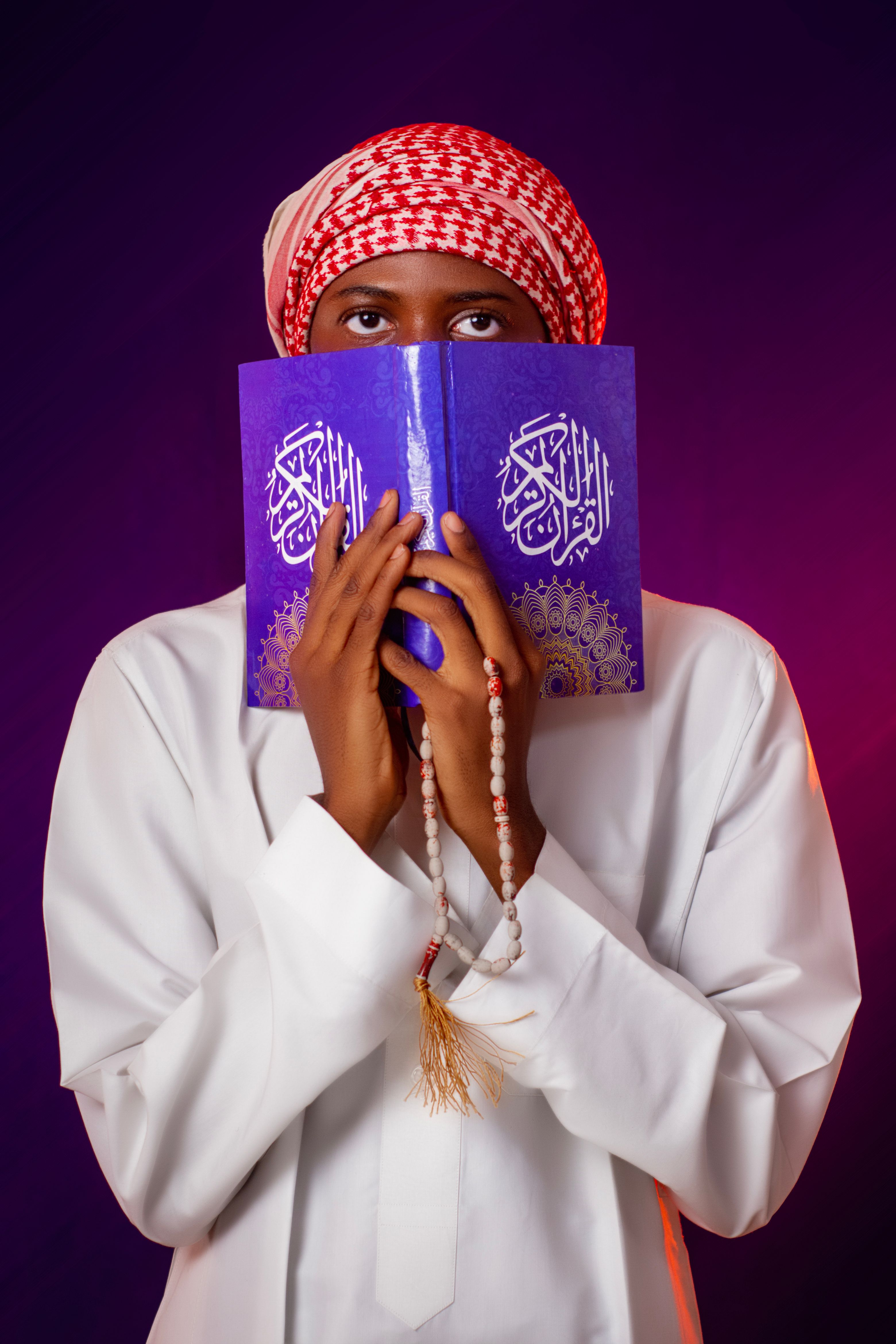 A man in traditional attire holds a Quran and prayer beads against a vibrant background.