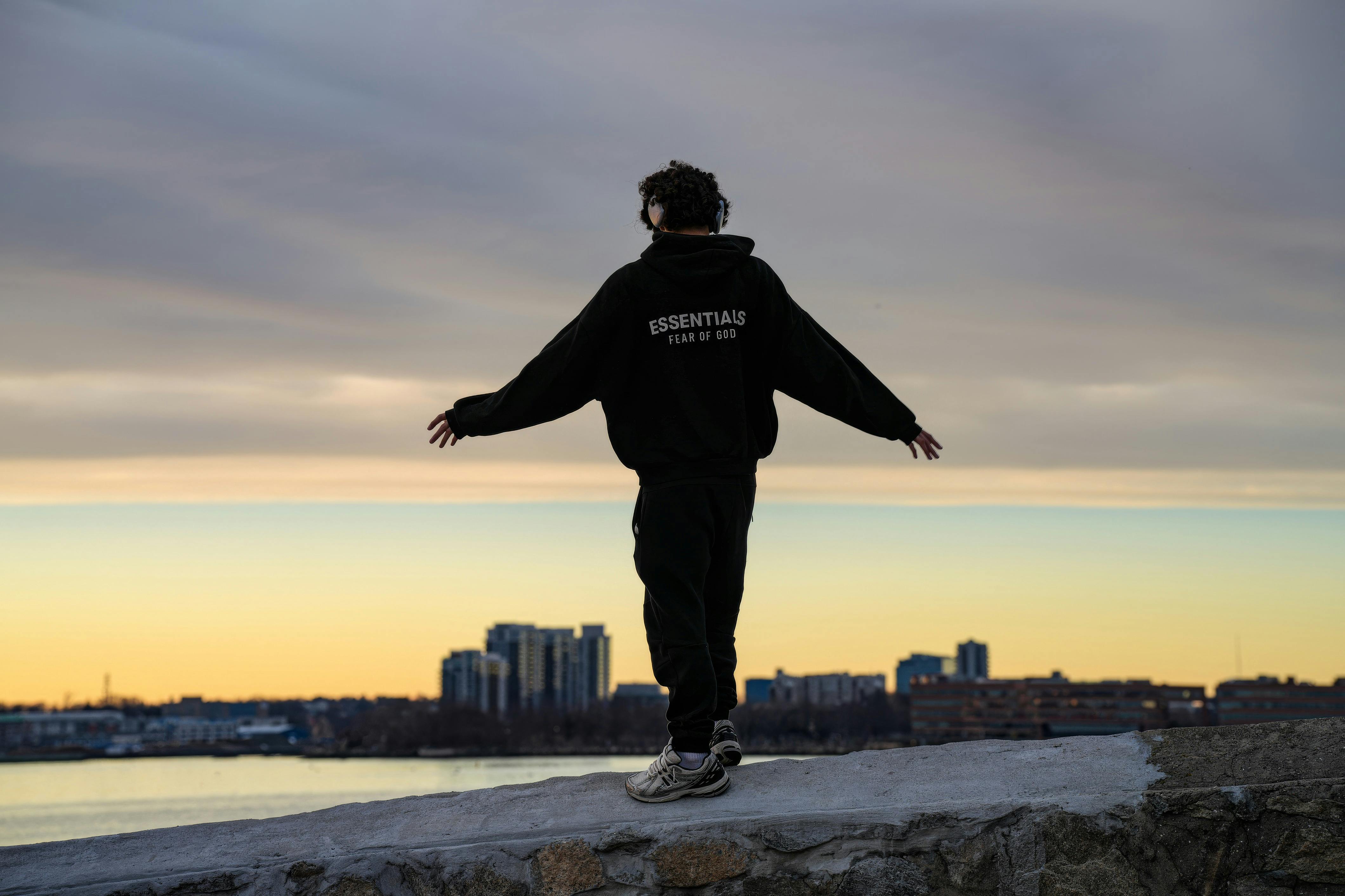 Gratis Un adolescente è in piedi su una sporgenza con sfondo urbano durante il tramonto, con le braccia tese. Foto a disposizione