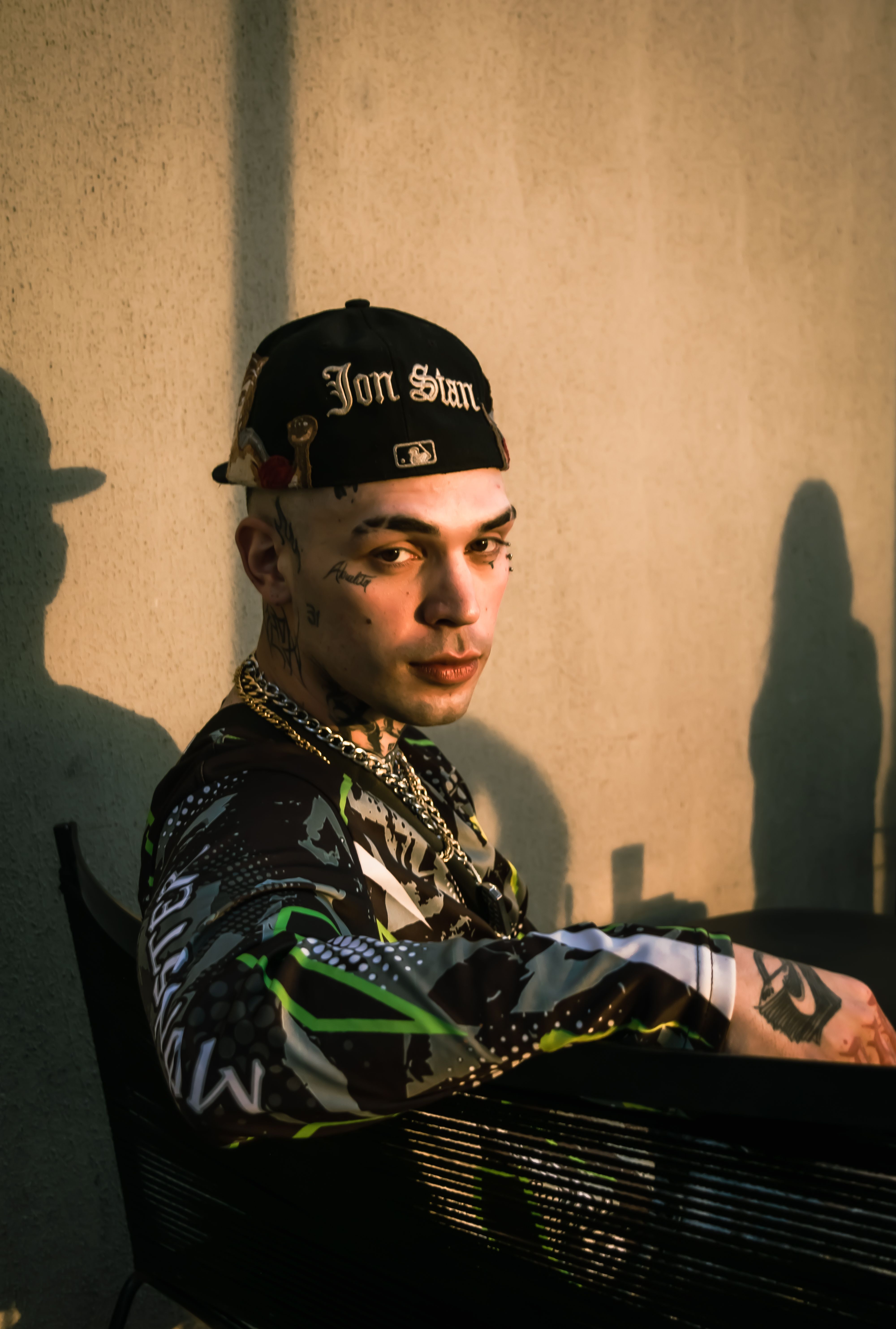Young man with tattoos sitting outdoors at sunset, wearing a cap and stylish clothes.