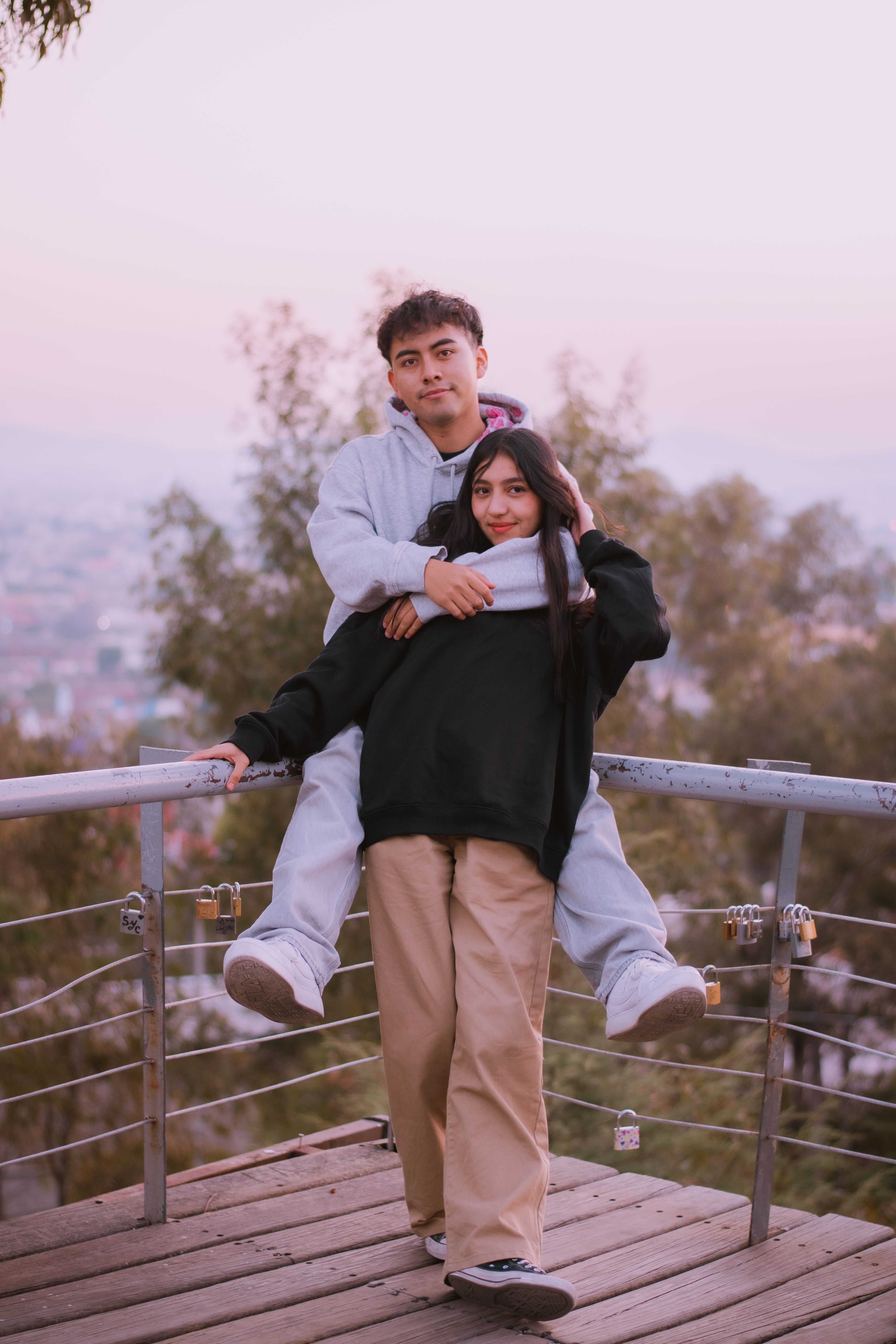 A young couple embraces on a scenic overlook, surrounded by trees and a pastel sky.