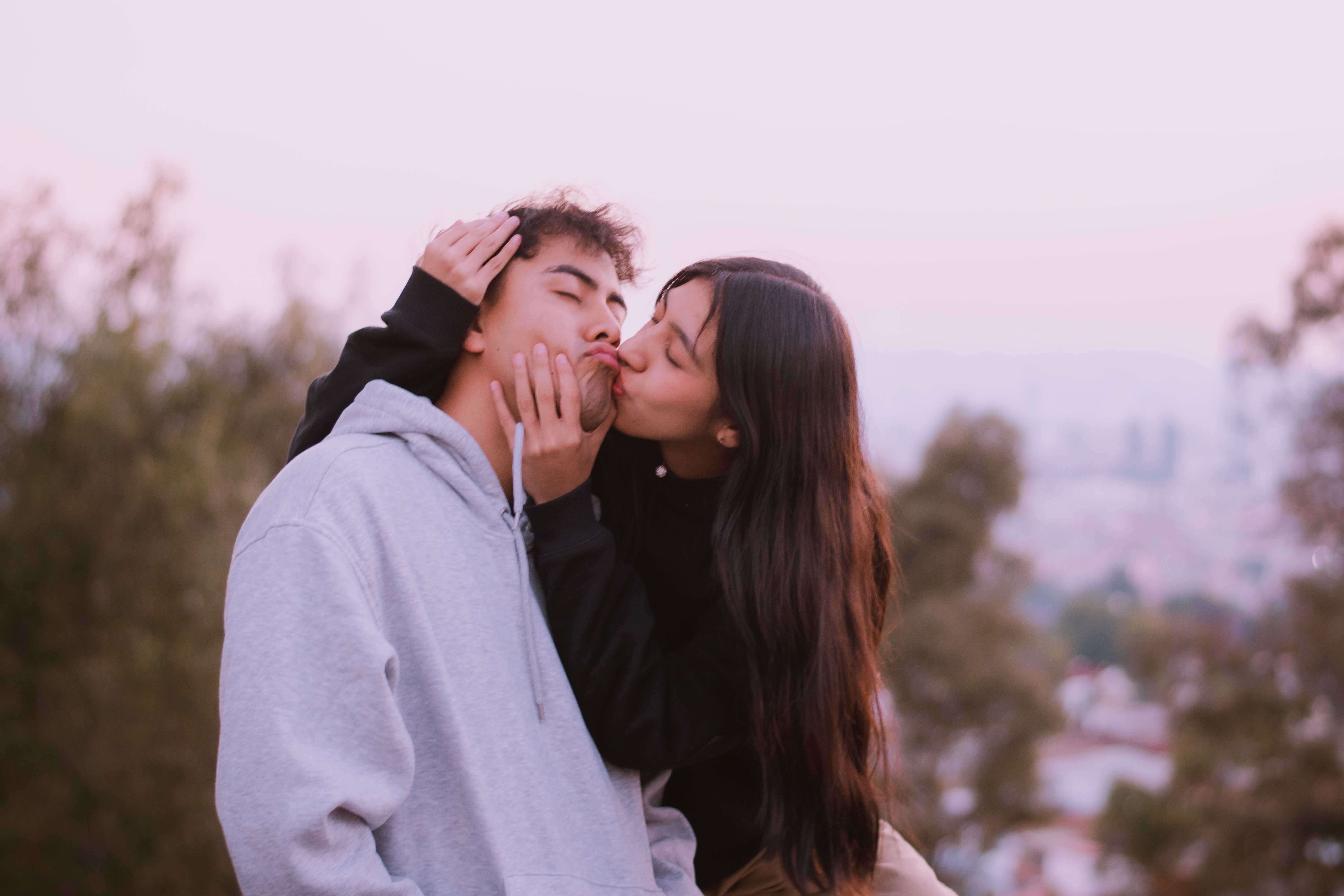 A young couple shares a tender kiss outdoors, immersed in a soft, romantic setting.