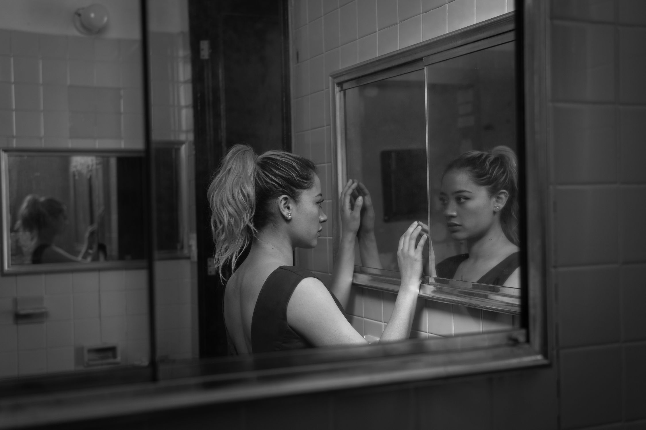 Fotografia em preto e branco de uma mulher olhando para o seu reflexo no espelho do banheiro.