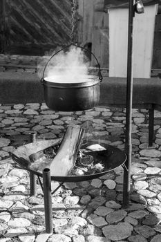 A black and white scene of a steaming pot hanging over an open fire on cobblestones.