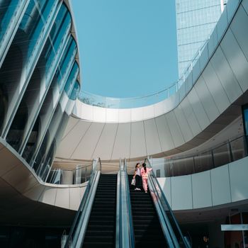 Two adults on an escalator within a sleek modern architectural structure under a clear blue sky.