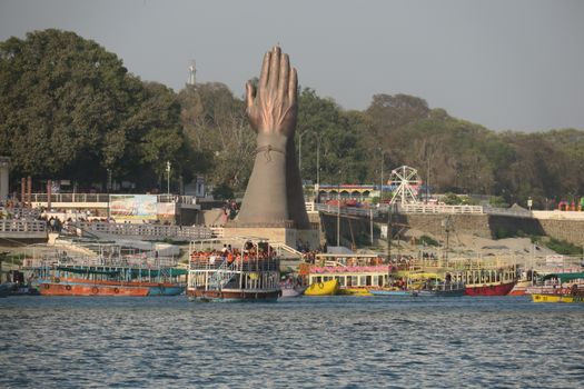 A large hand statue near a bustling riverbank with colorful tour boats in the foreground.