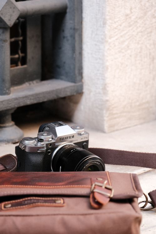 Free Modern Fujifilm camera with a leather bag placed on an outdoor stone surface. Photographic gear setup. Stock Photo
