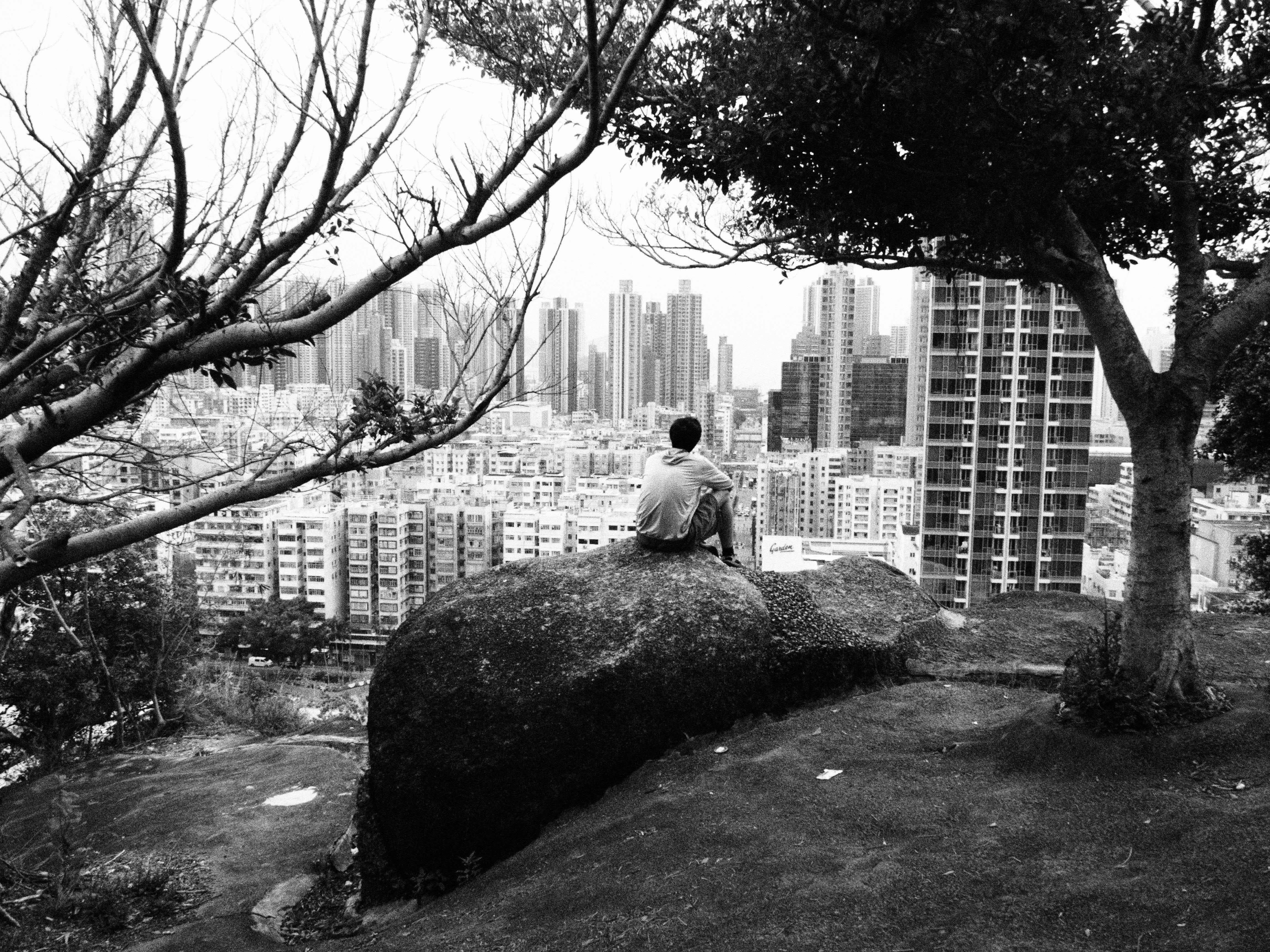 A person sits on a rock overlooking the urban skyline of Hong Kong, framed by trees.