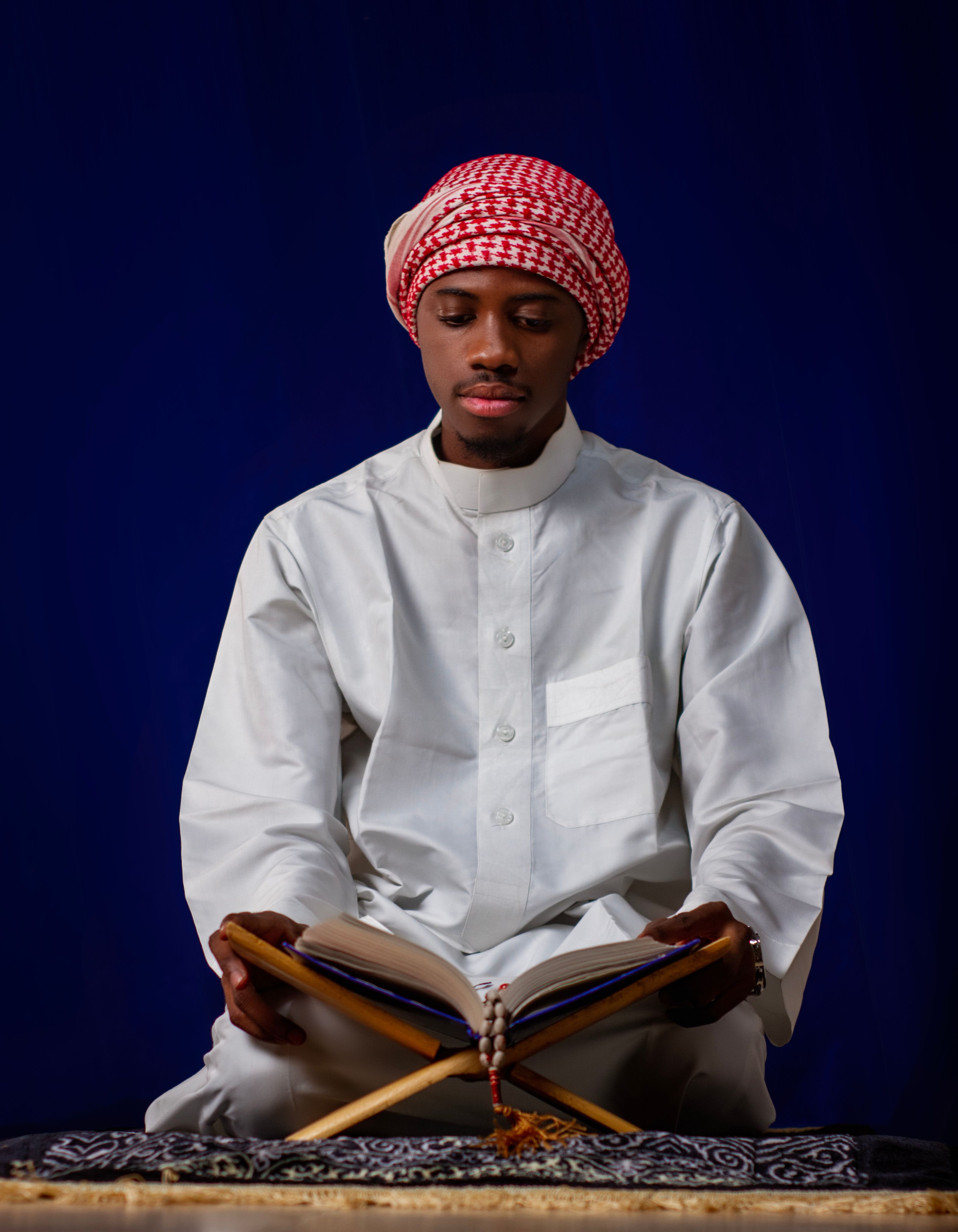 Middle-Eastern man in white attire reading the Quran, set against a rich blue background.