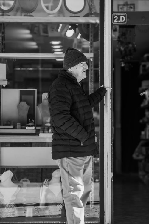 Free Black and white photo of an elderly man in a winter coat standing outside a shop. Stock Photo