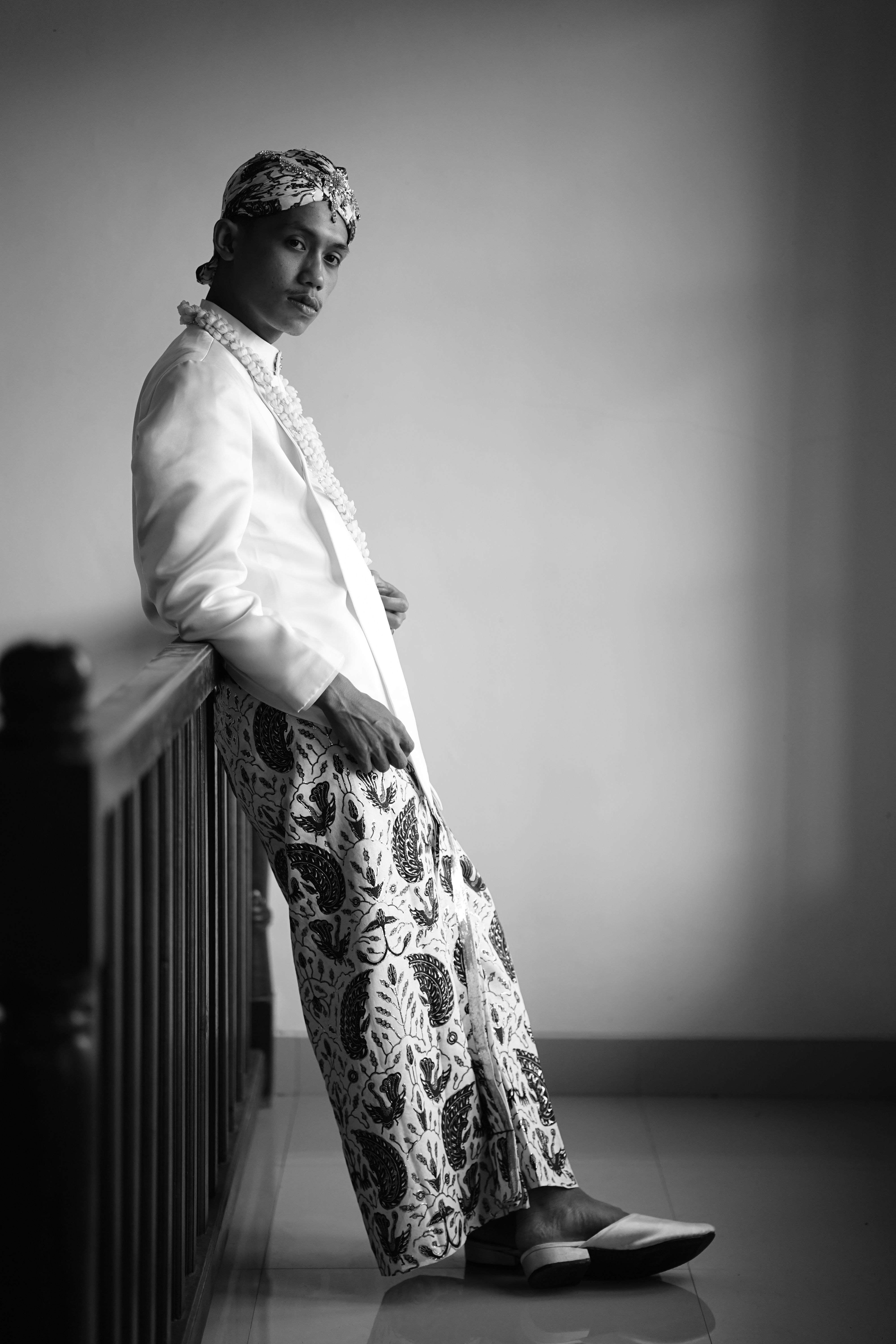 Man in traditional Indonesian clothing, leaning against railing, captured in black and white.