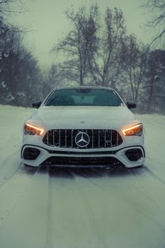 Front view of a luxury car on a snowy road, headlights glowing in winter setting.