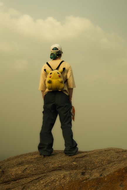 A young adult stands on a mountaintop with a Pikachu backpack, looking at the horizon.