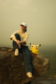 Person with a hat sits on a rock accompanied by a Pikachu plush toy, surrounded by misty landscape.